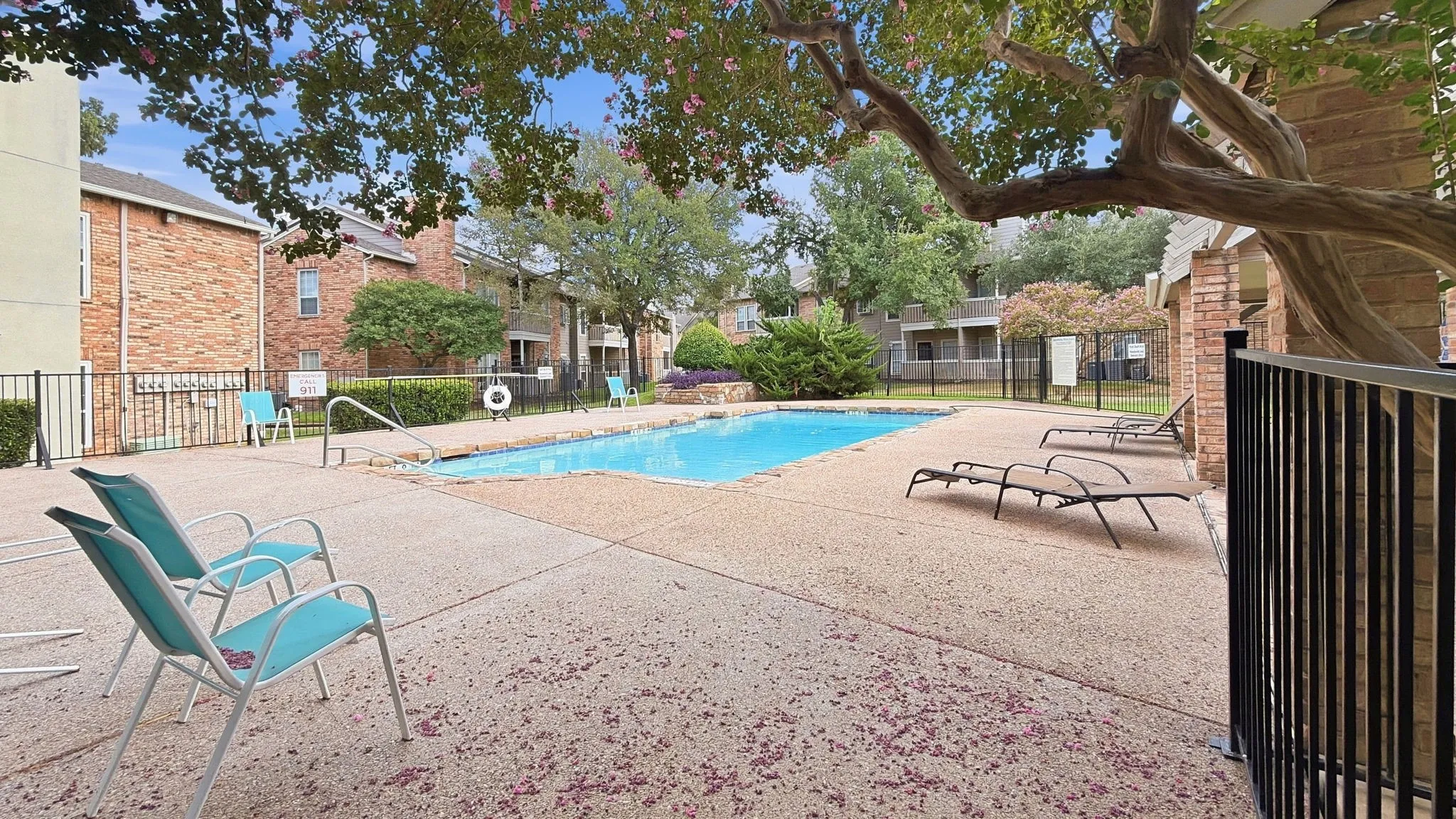 Outdoor swimming pool with concrete decking, surrounding brick buildings, and mature trees