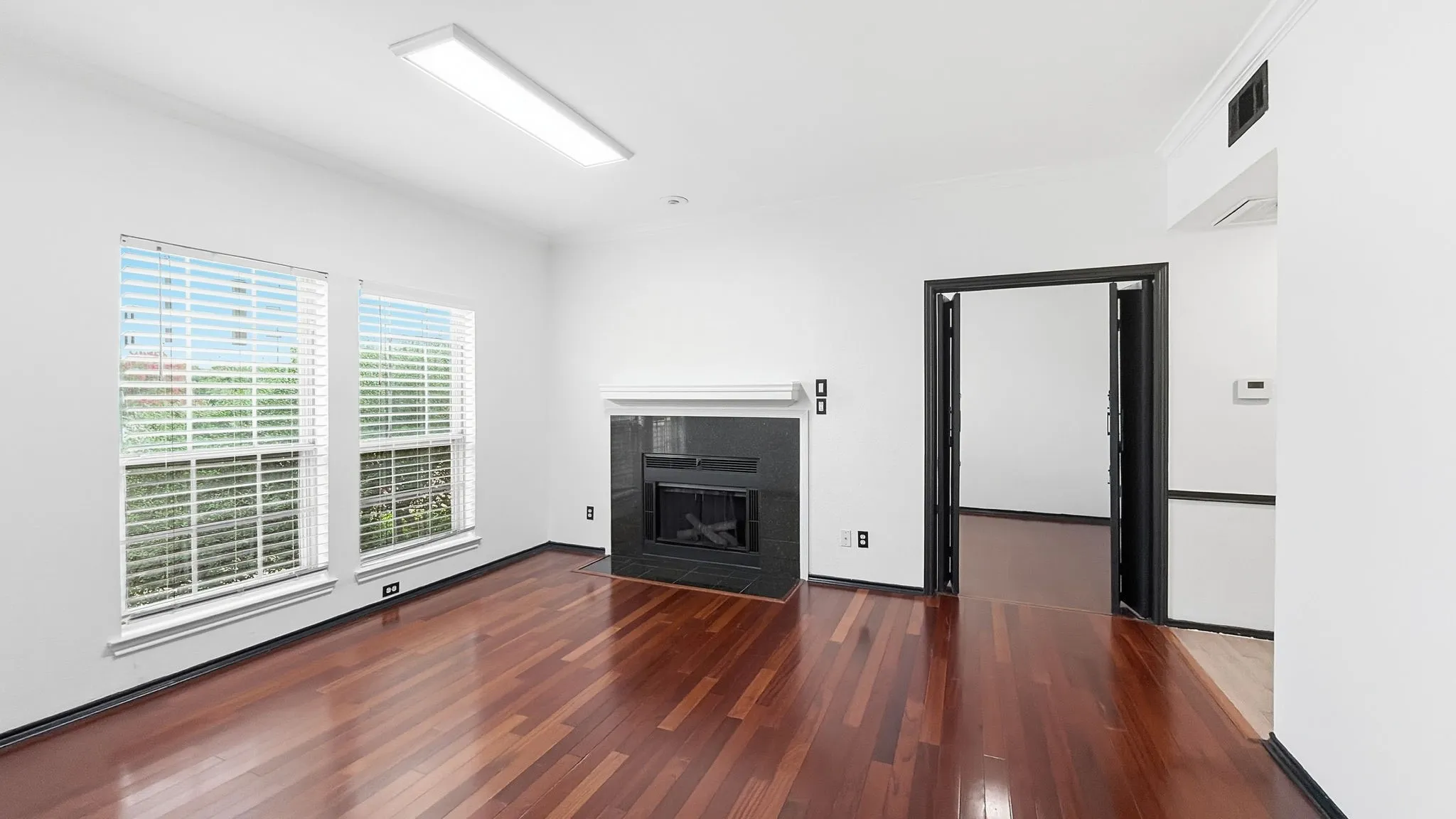 Rich wood-finish flooring, a fireplace with a dark-toned surround and white mantel, dual windows with blinds, and a recessed ceiling light fixture