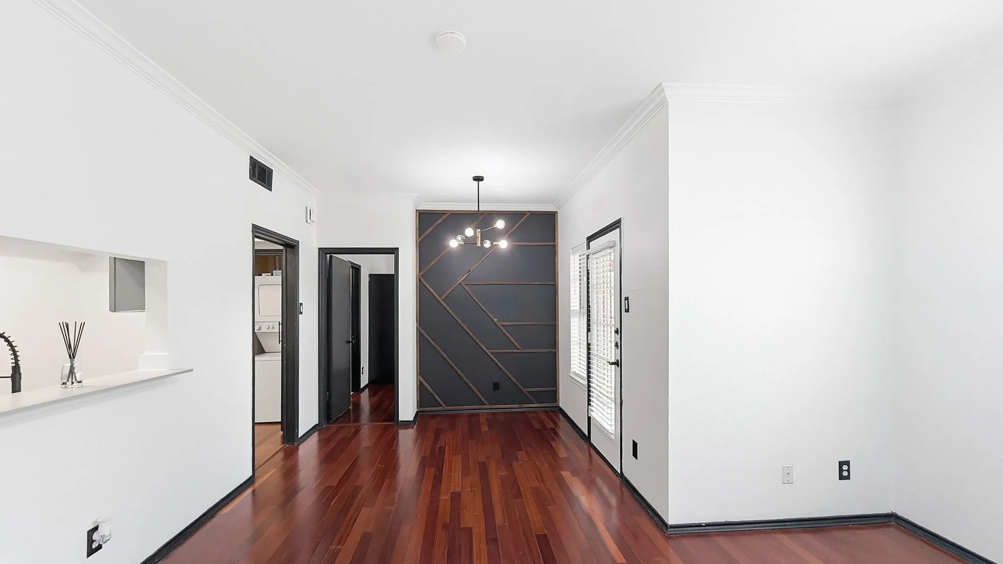 Rich wood-finish flooring, accent wall with geometric paneling, modern pendant light fixture, and crown molding