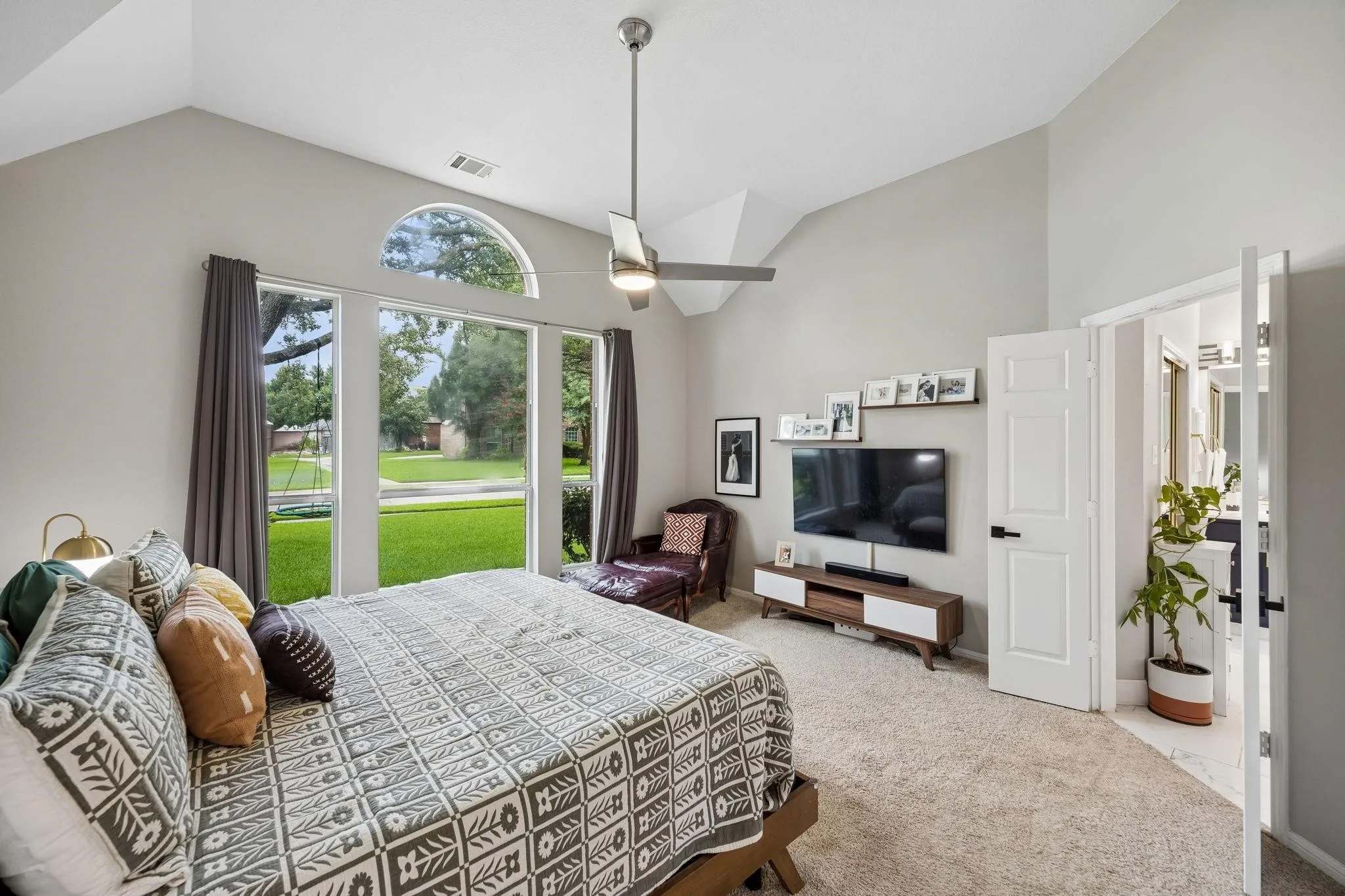 Vaulted ceiling with a ceiling fan, arched transom window, large windows with exterior landscaping, carpet flooring, and a paneled door