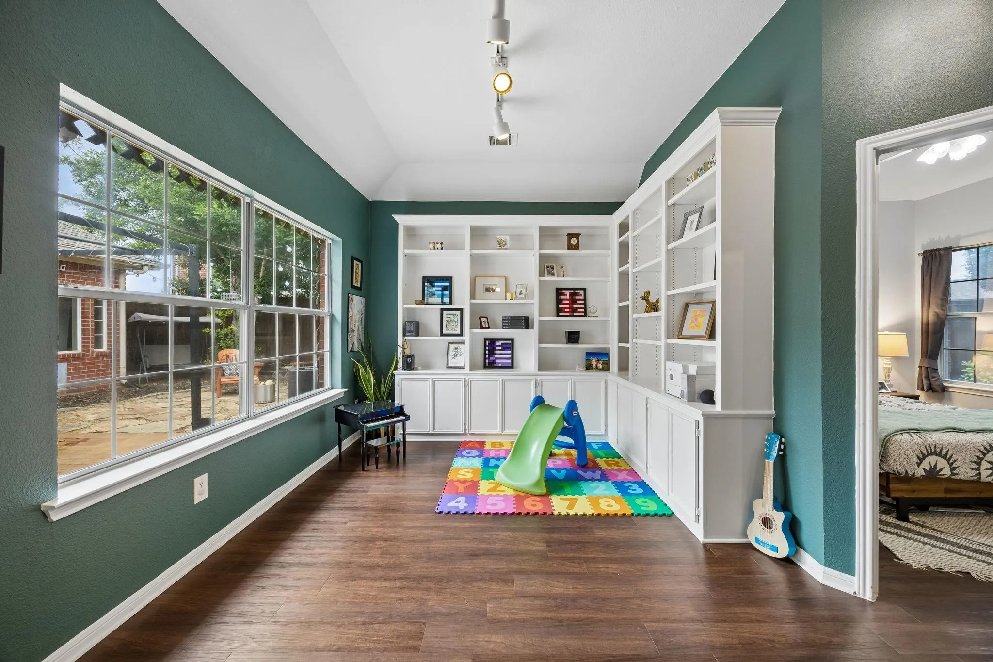 Vaulted ceiling with track lighting, wood-finish flooring, extensive built-in shelving, multiple windows, and a doorway opening to another room