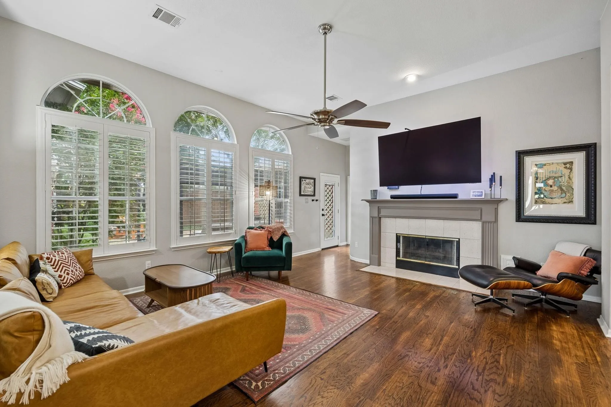 Arched windows with plantation shutters, wood-finish flooring, vaulted ceilings, a ceiling fan, and a fireplace with a tile surround and mantel