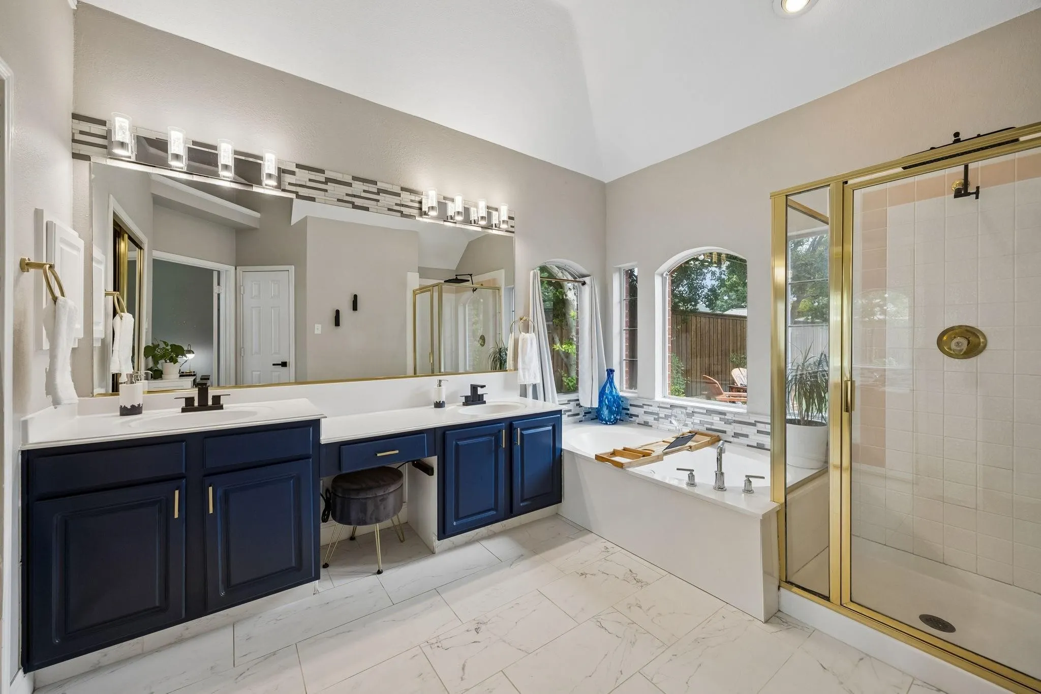 Vaulted ceiling over a bathroom featuring a large built-in tub, a glass-enclosed shower, dual vanities with dark cabinetry, and white tile flooring