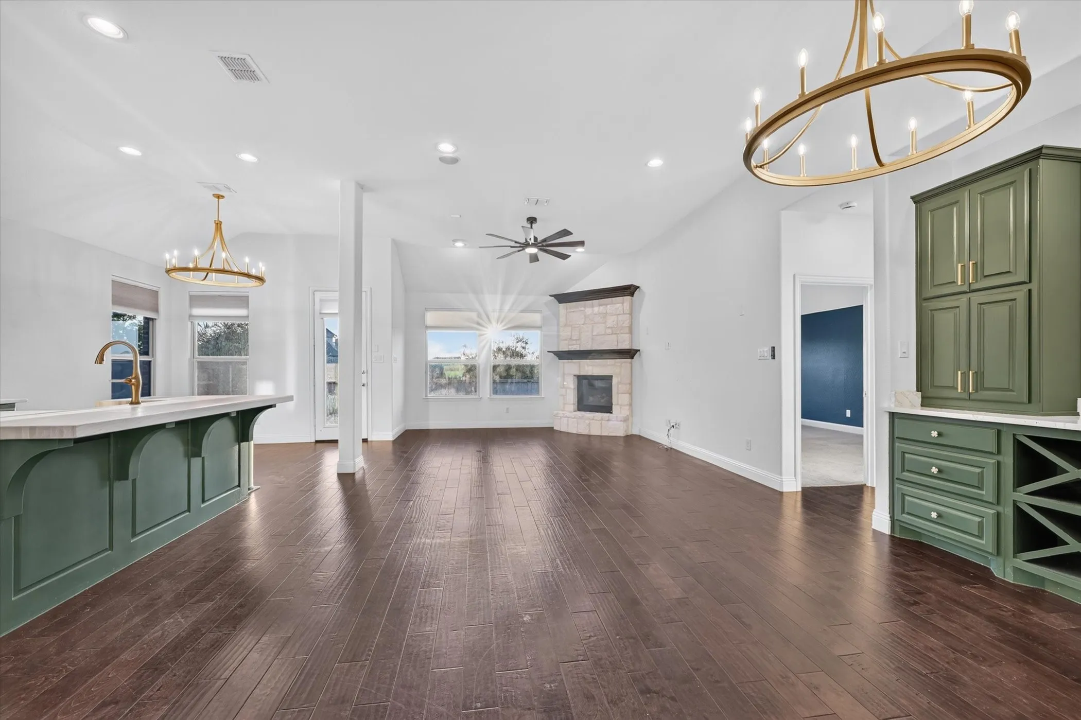 Vaulted ceiling with recessed lighting, wood-finish flooring, stone-clad fireplace, built-in cabinetry with wine storage, and a kitchen island with a gold-tone faucet