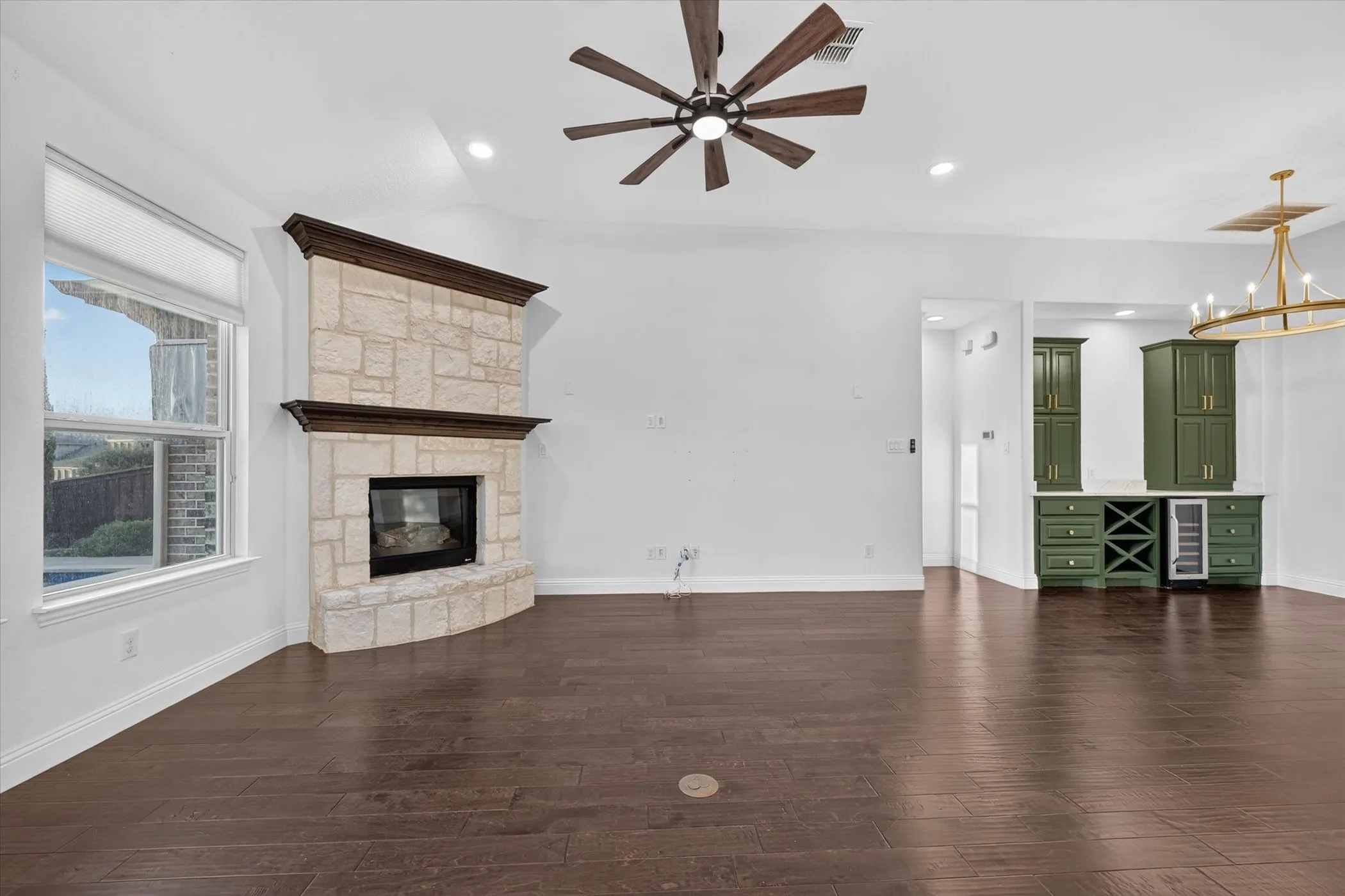 Stone-faced fireplace with a dark wood mantel, rich wood-finish flooring, built-in wet bar with green cabinetry, and a ceiling fan with integrated lighting