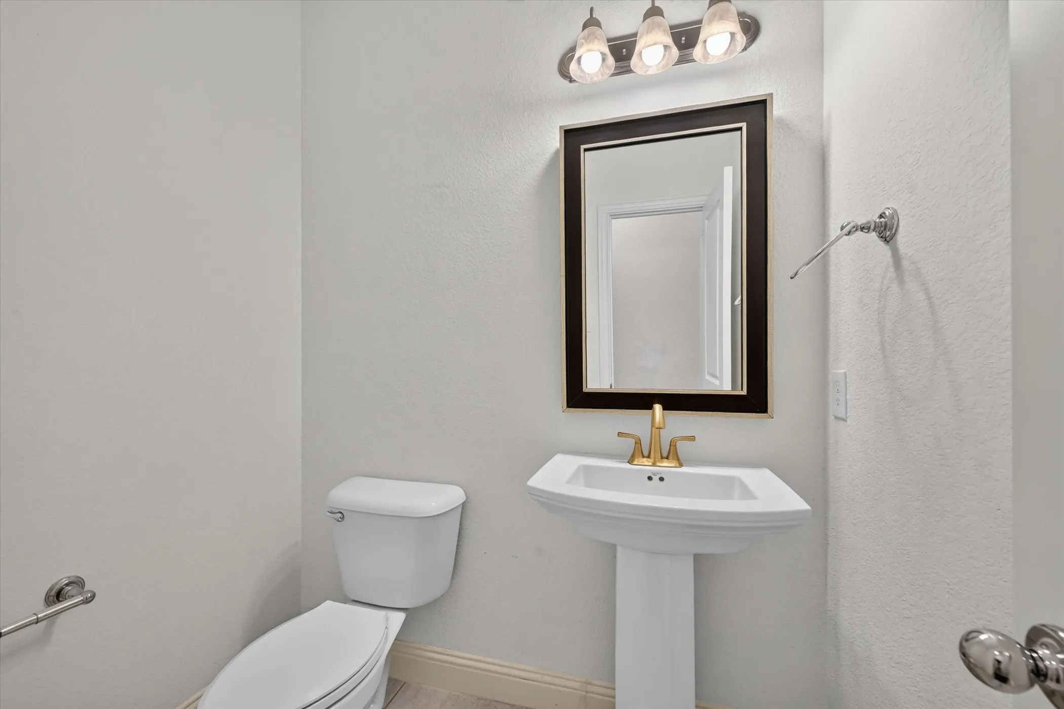 Pedestal sink with a gold-tone faucet, framed mirror, and a three-light vanity fixture