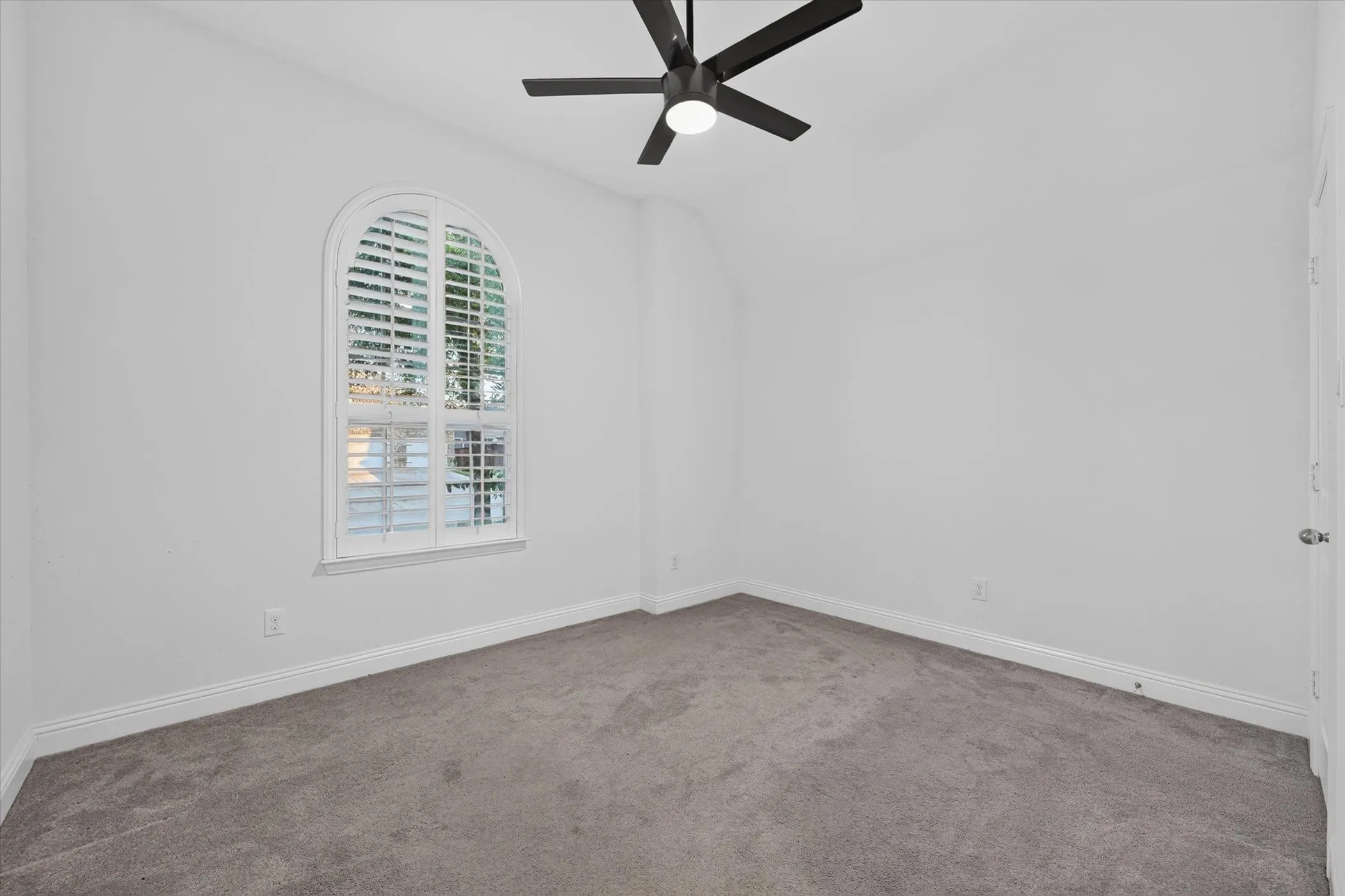 Arched window with plantation shutters, light gray carpet flooring, white walls, and a ceiling fan with integrated lighting
