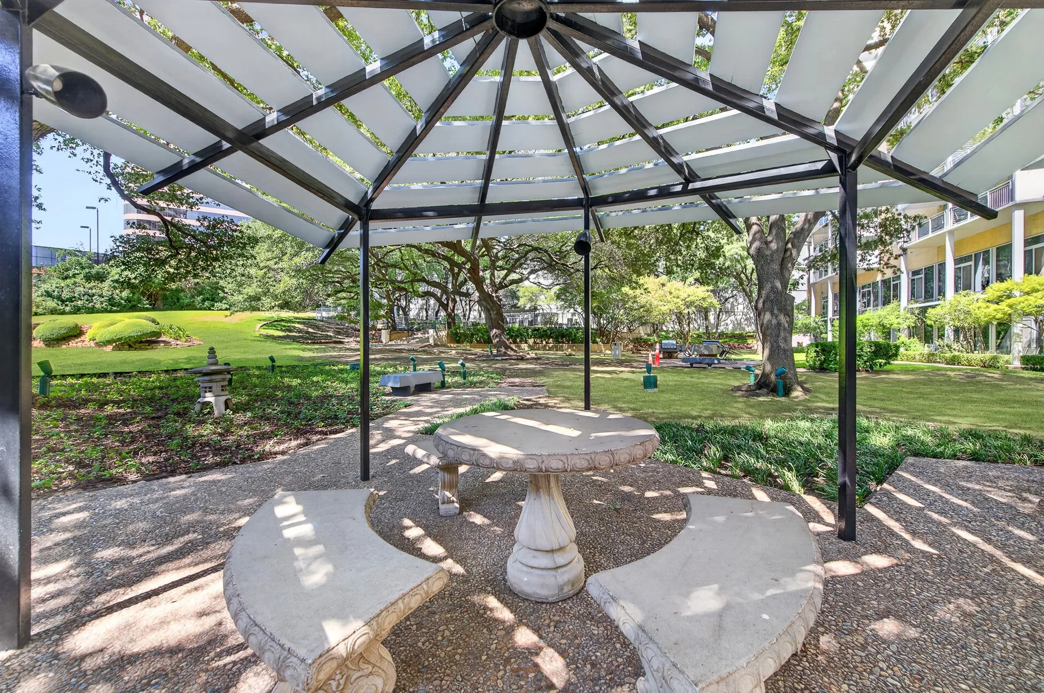 Shaded outdoor seating area with a stone table and benches, set on a gravel surface