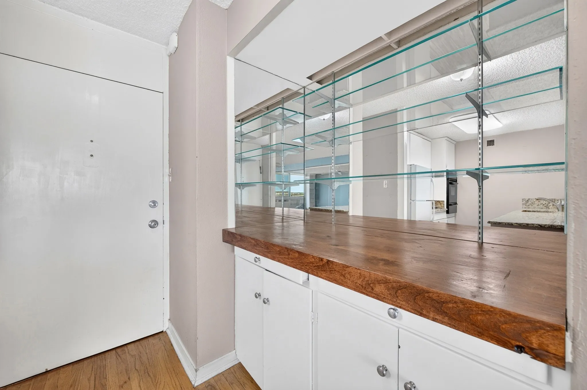 Wood flooring, white entry door, built-in cabinetry with white doors, wood countertop, and glass shelving
