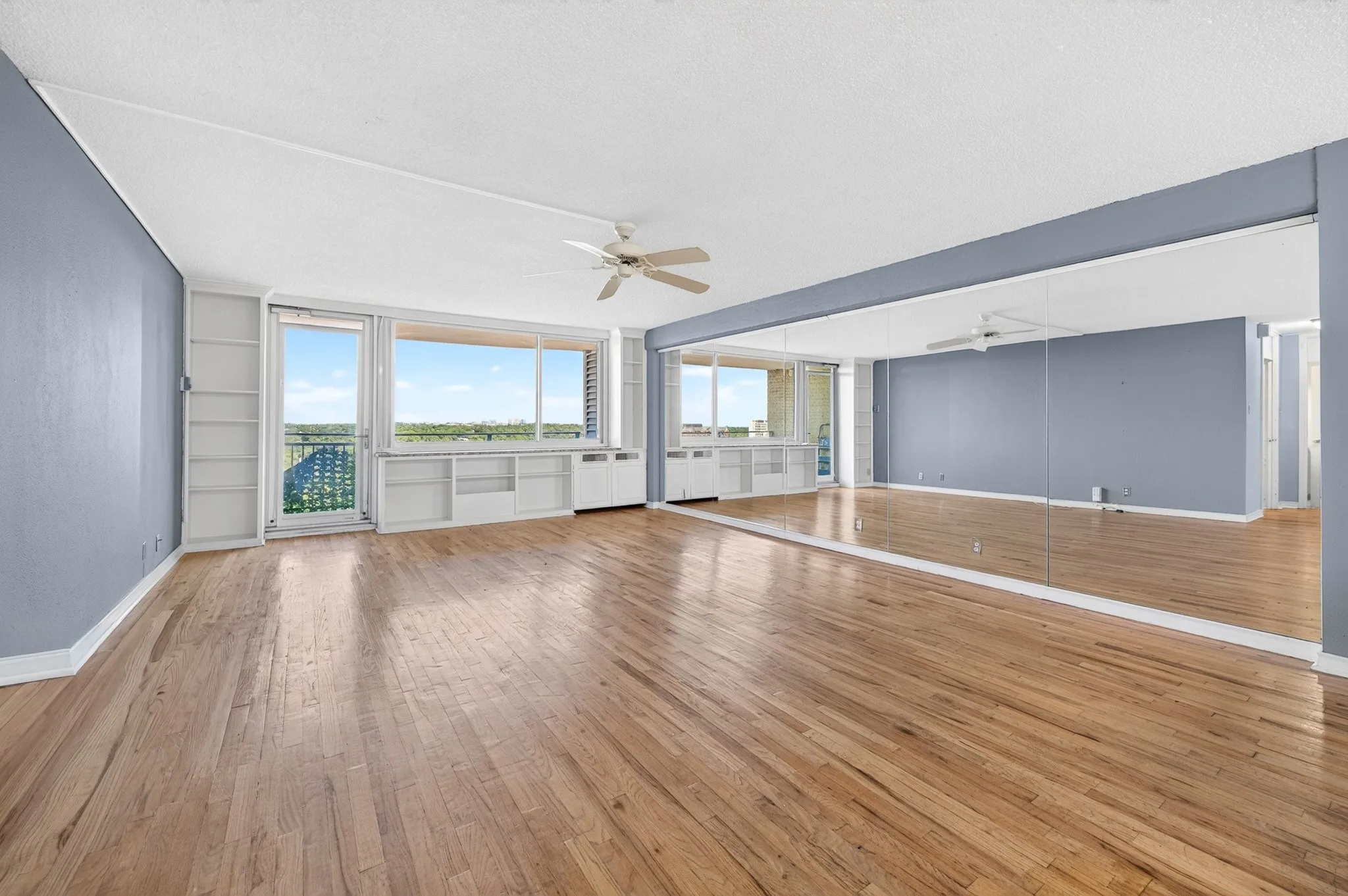 Expansive living area with wood flooring, full-wall mirror, built-in shelving, and a ceiling fan