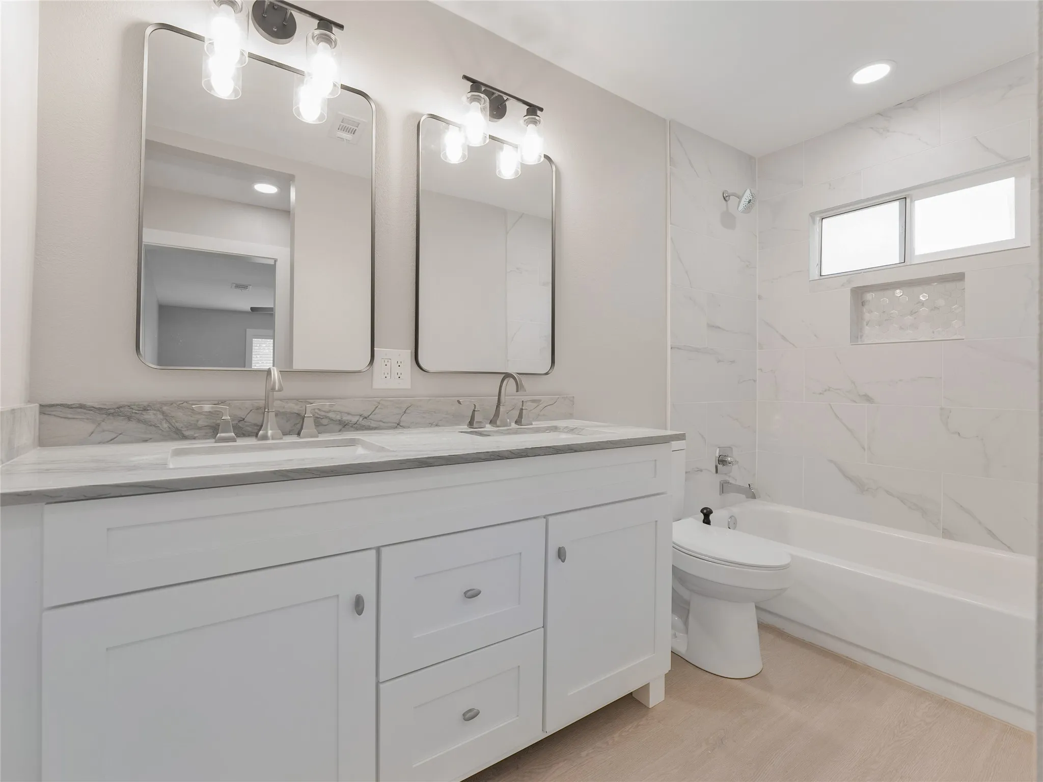 Dual vanity with undermount sinks and light-toned countertops, individual rectangular mirrors with sconce lighting, integrated bathtub, and large format wall tiling