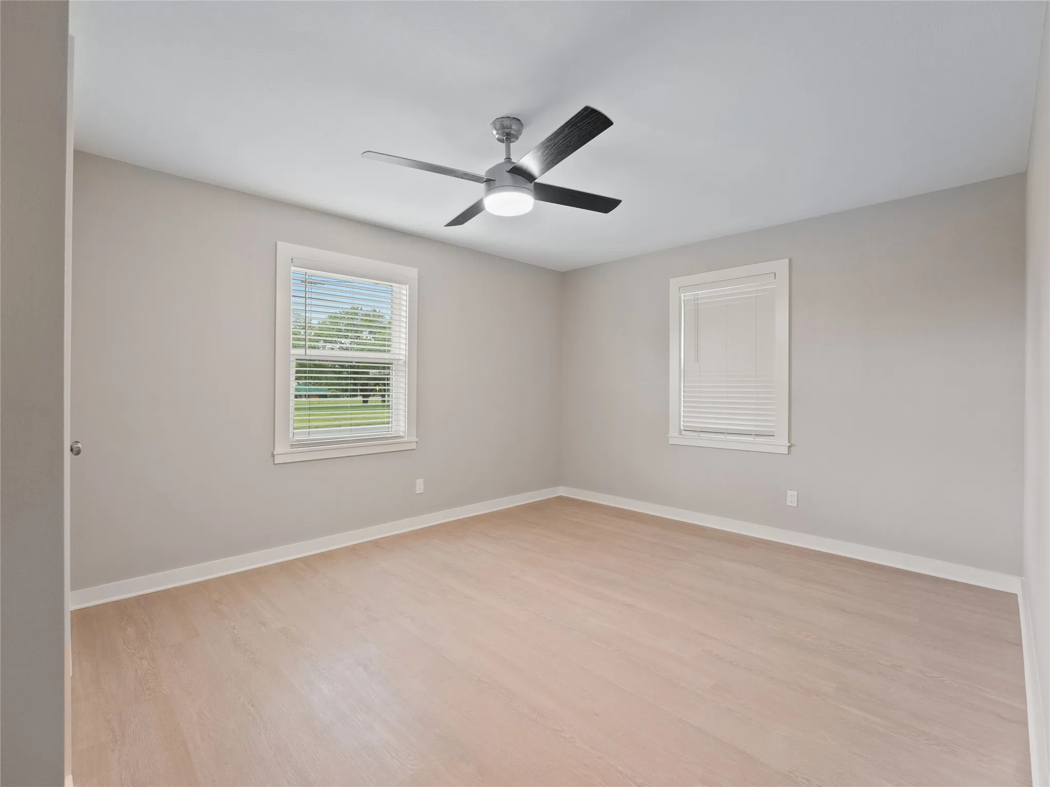 Light wood-finish flooring, two windows with white blinds, and a ceiling fan with integrated lighting