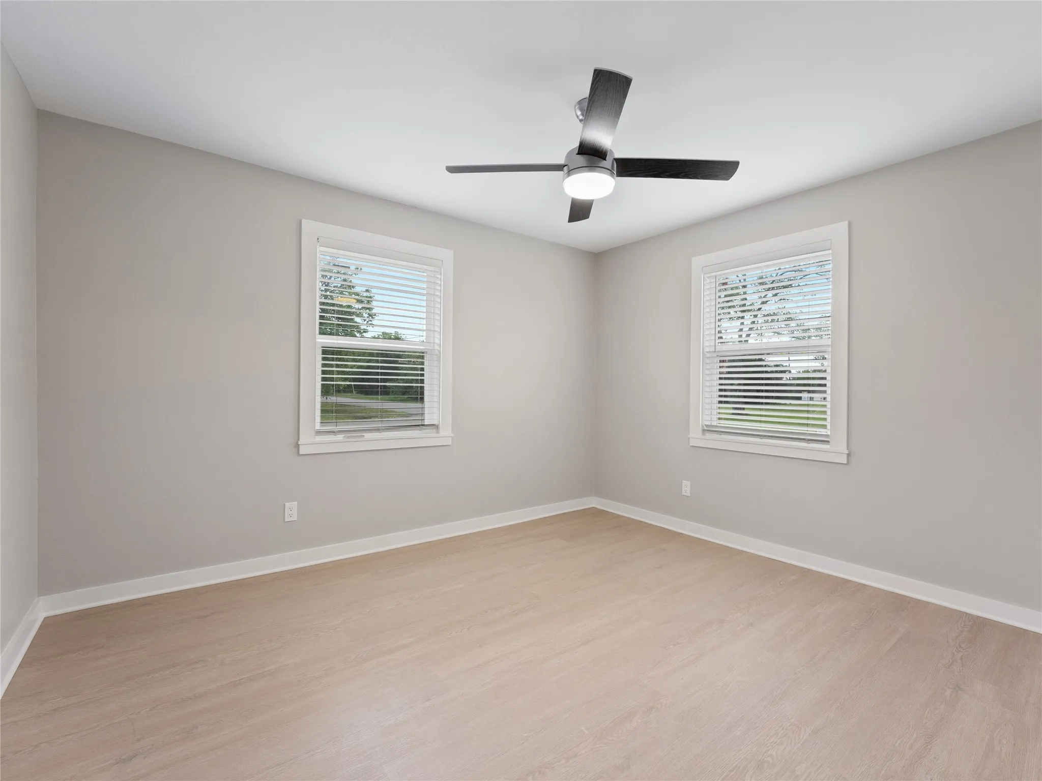 Light-toned wood-finish flooring, neutral wall paint, two windows, and a ceiling fan with integrated lighting