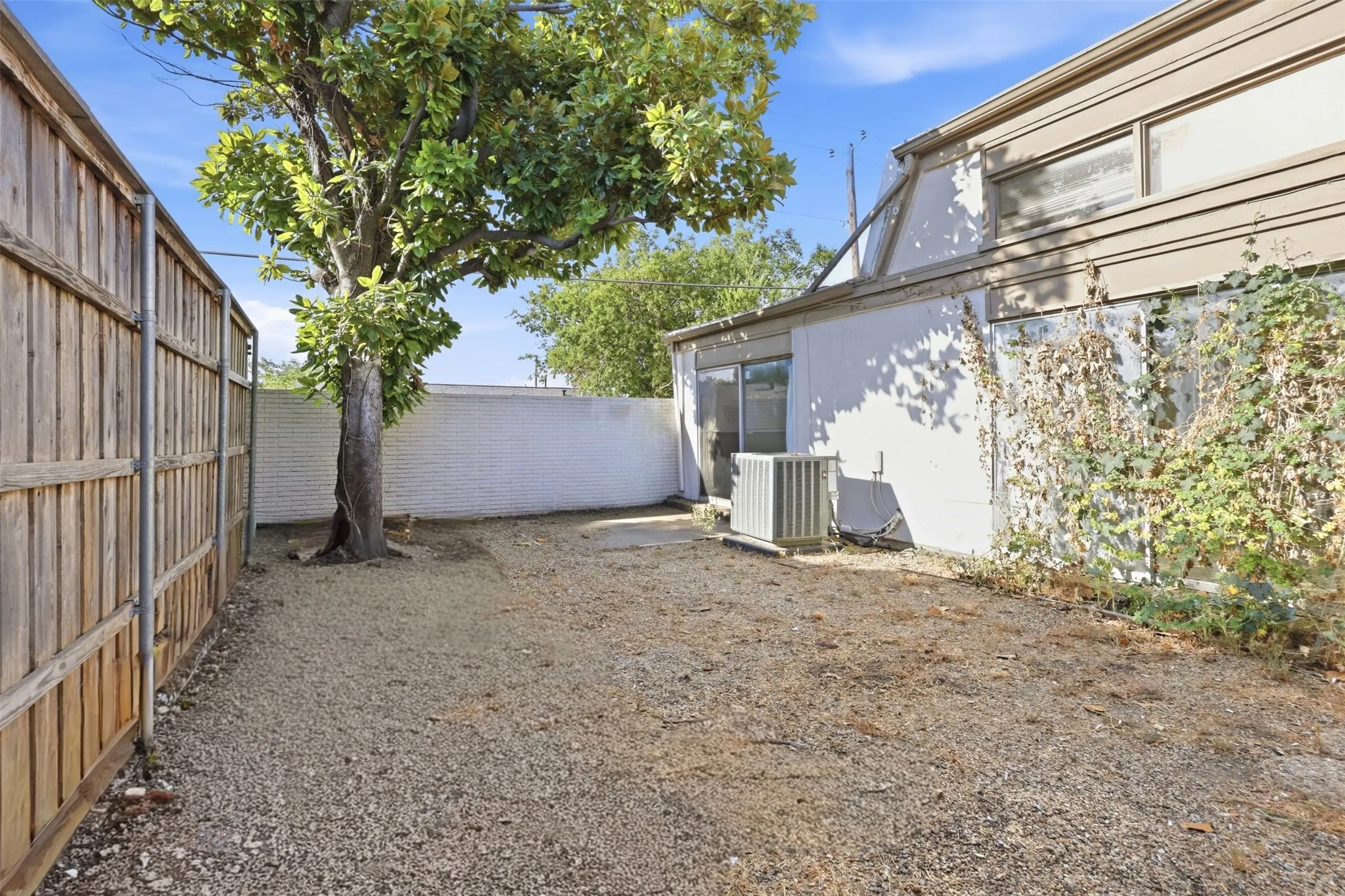 Enclosed exterior space featuring a mature tree, gravel ground covering, wood privacy fence, and a white painted brick wall