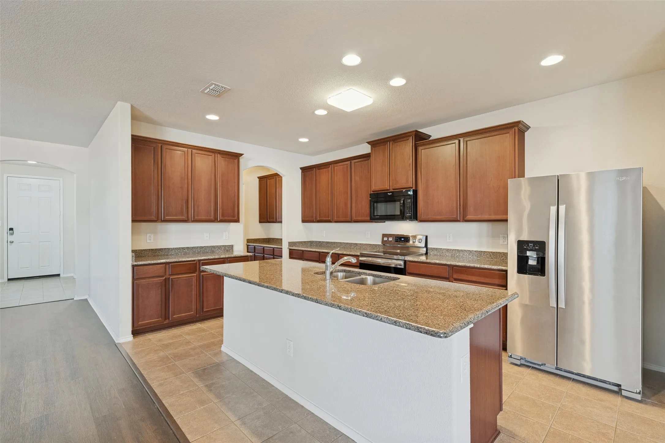 Kitchen featuring wood cabinetry, a center island with sink, granite-patterned countertops, and tile flooring