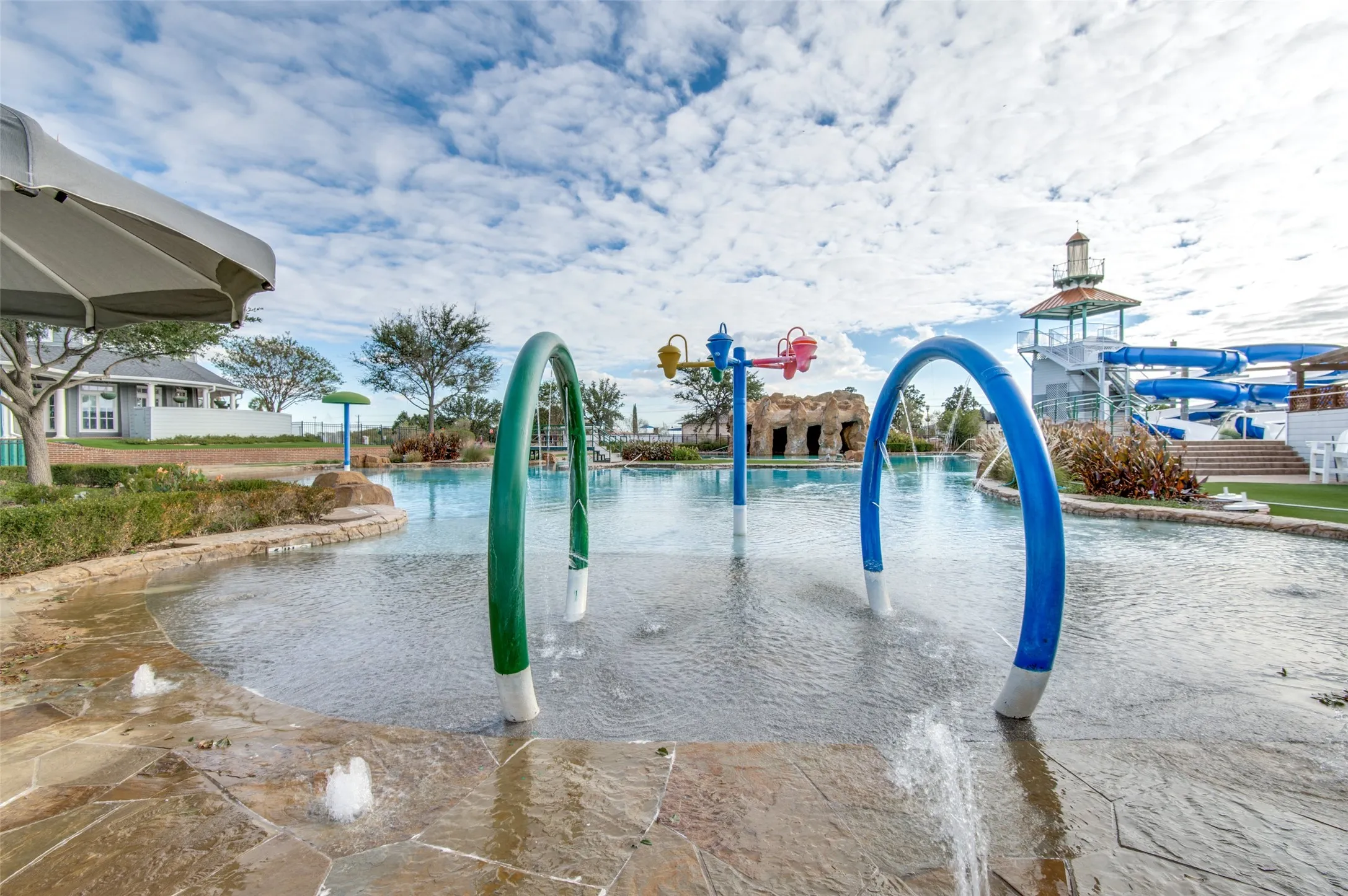 Stone-edged splash pad with water features, adjacent to a resort-style pool with a lighthouse-inspired water slide structure and a nearby building with exterior siding