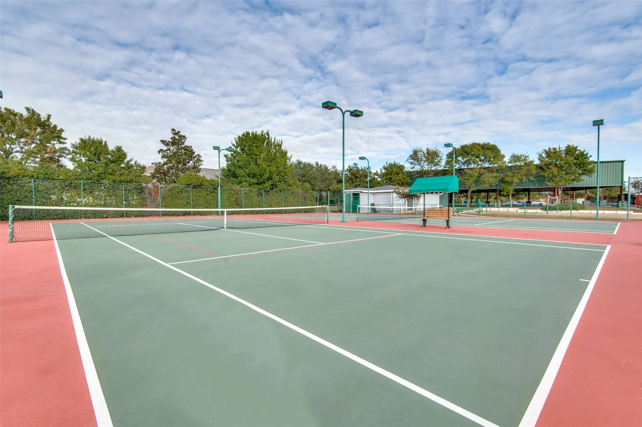 Tennis court with hard surface and red and green playing areas, perimeter fencing, and overhead lighting