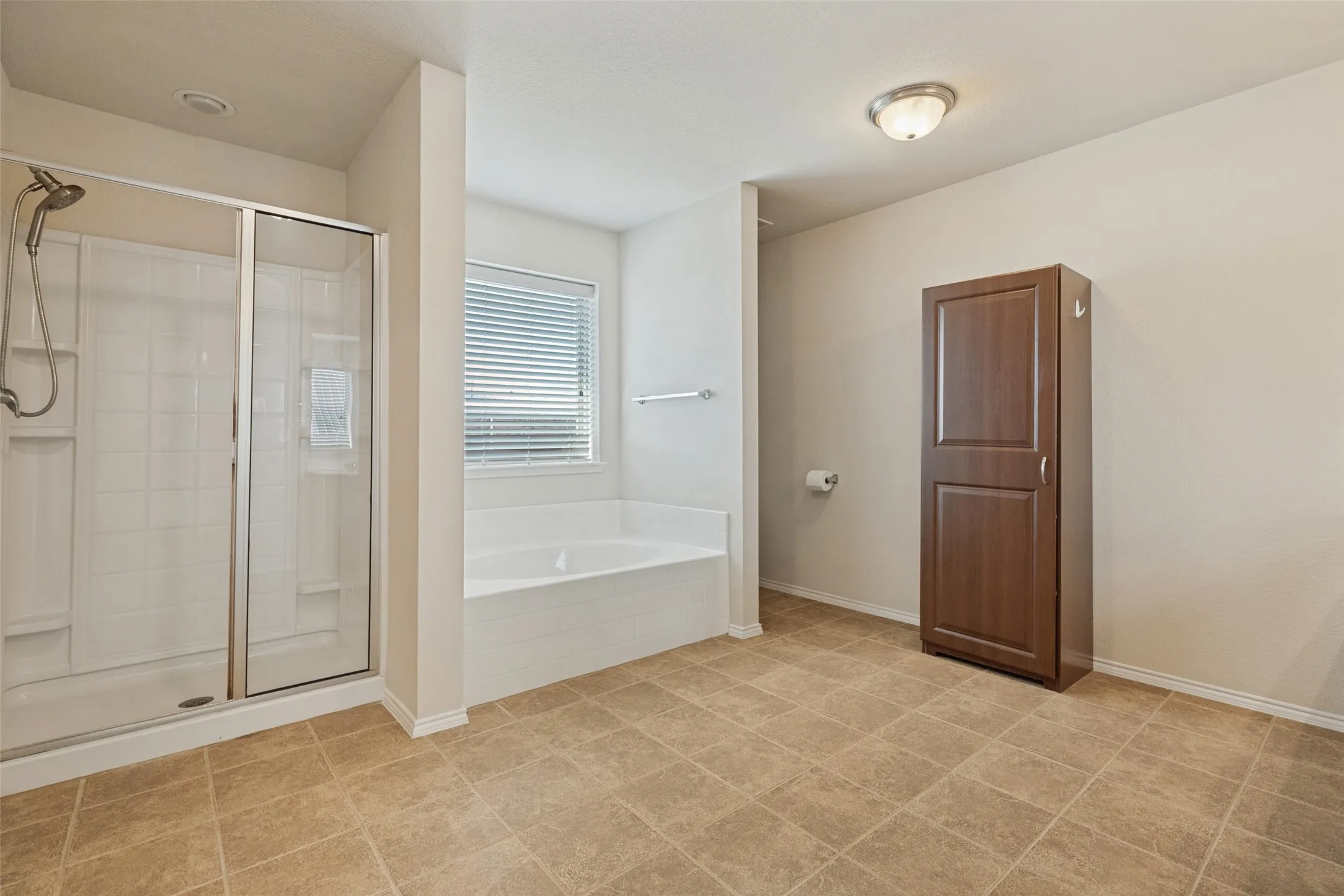 Tiled flooring, built-in soaking tub, separate shower with glass enclosure, window with blinds, and a tall wood-finish storage cabinet