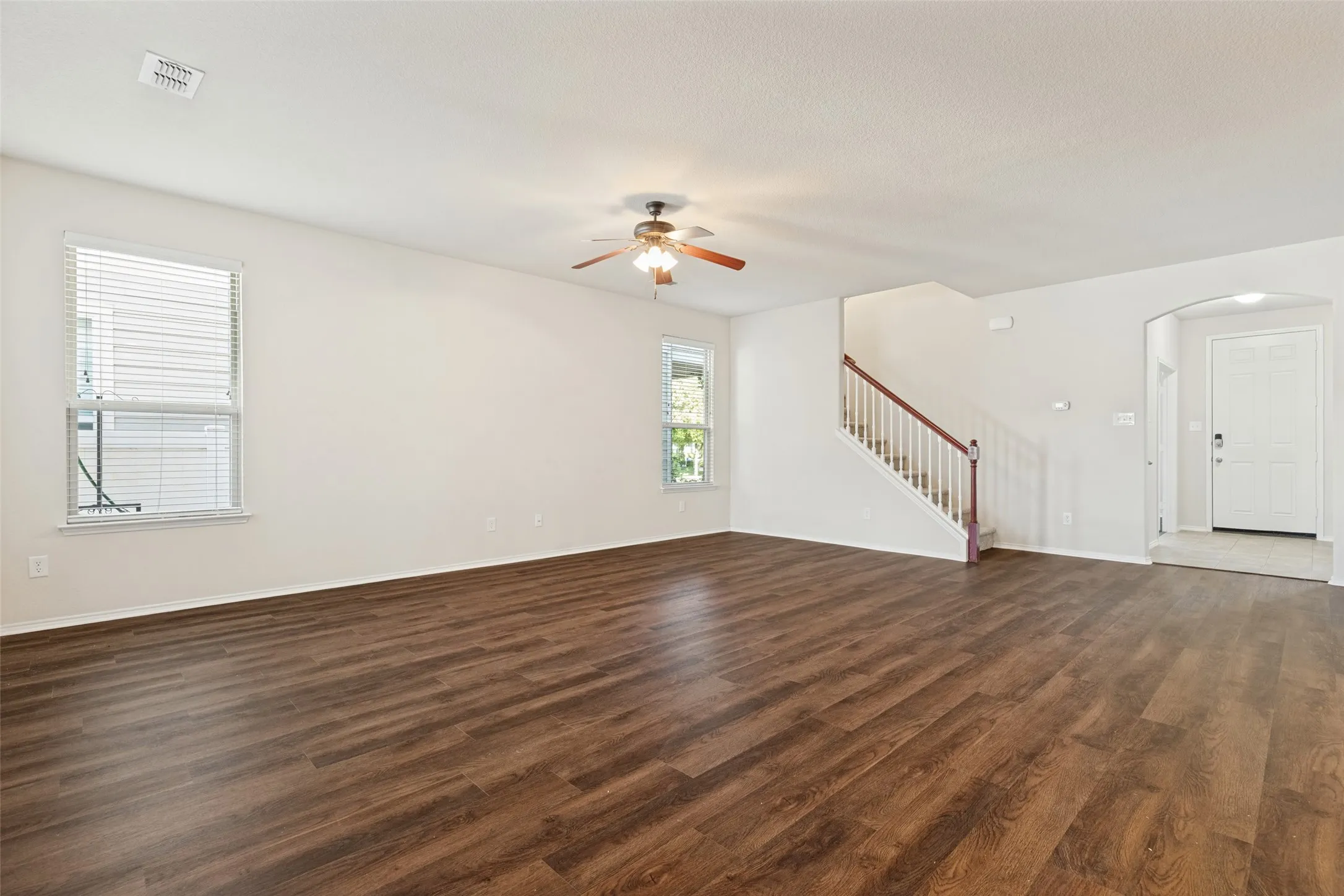 Spacious living area featuring wood-finish flooring, multiple windows with blinds, a ceiling fan, and a staircase with a wooden handrail