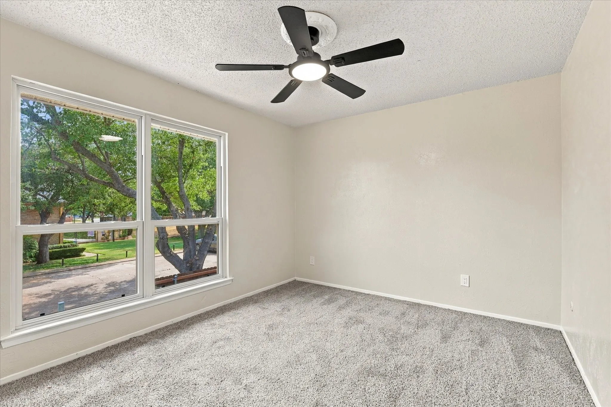 Expansive window offering natural illumination, equipped with a ceiling fan with integrated lighting, soft grey carpeting, and neutral wall tones