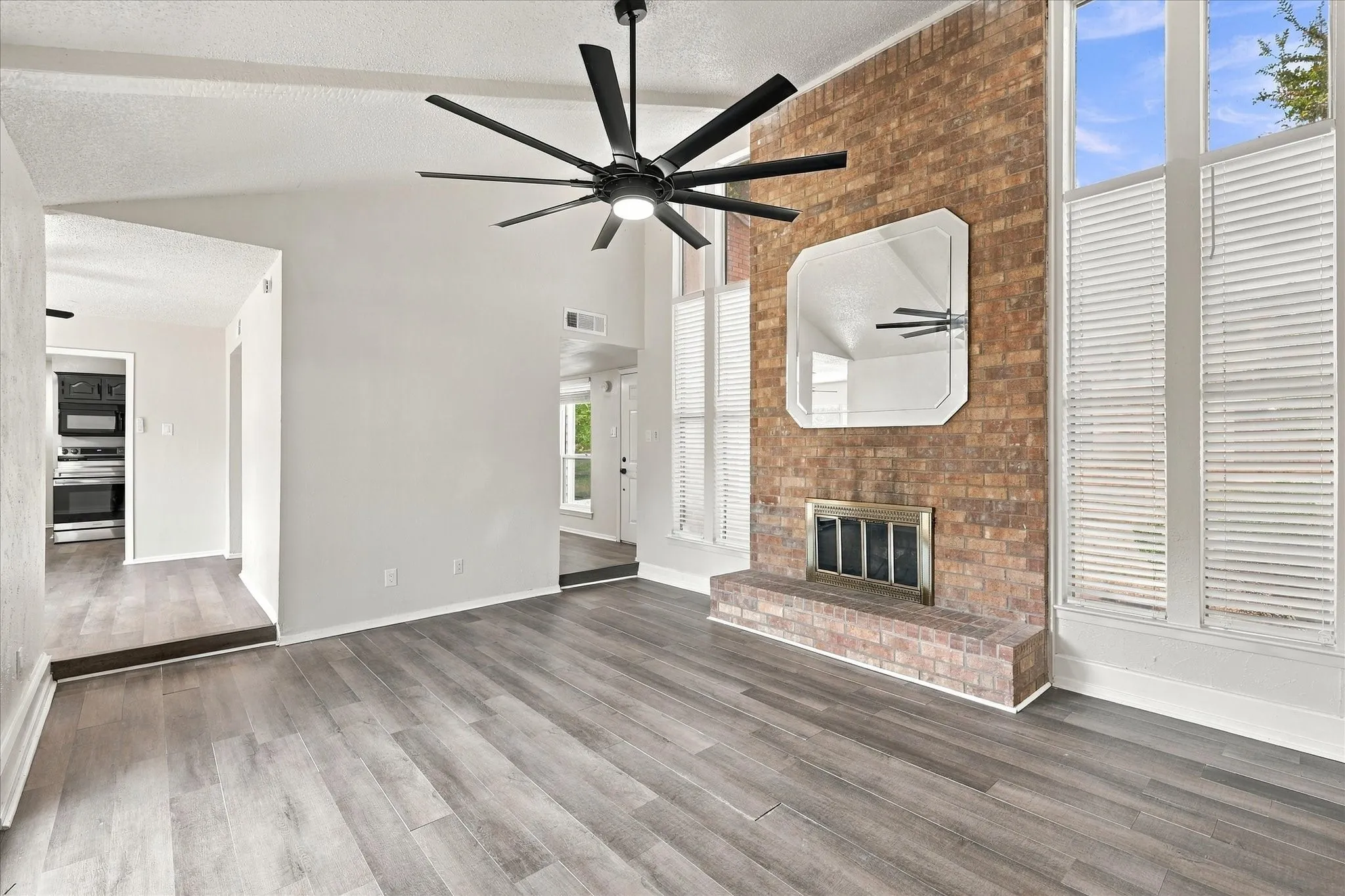 Vaulted ceiling featuring a contemporary ceiling fan, brick fireplace with a raised hearth, wood-finish flooring, and tall windows with interior shutters