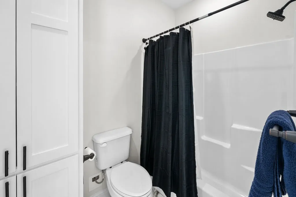 White paneled cabinetry with dark hardware, a white toilet, and a shower with a white surround and dark-finish showerhead
