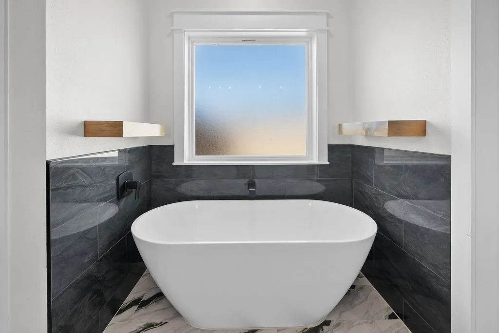 Freestanding soaking tub with floor-mounted faucet, dark stone tile surround, and window with white trim