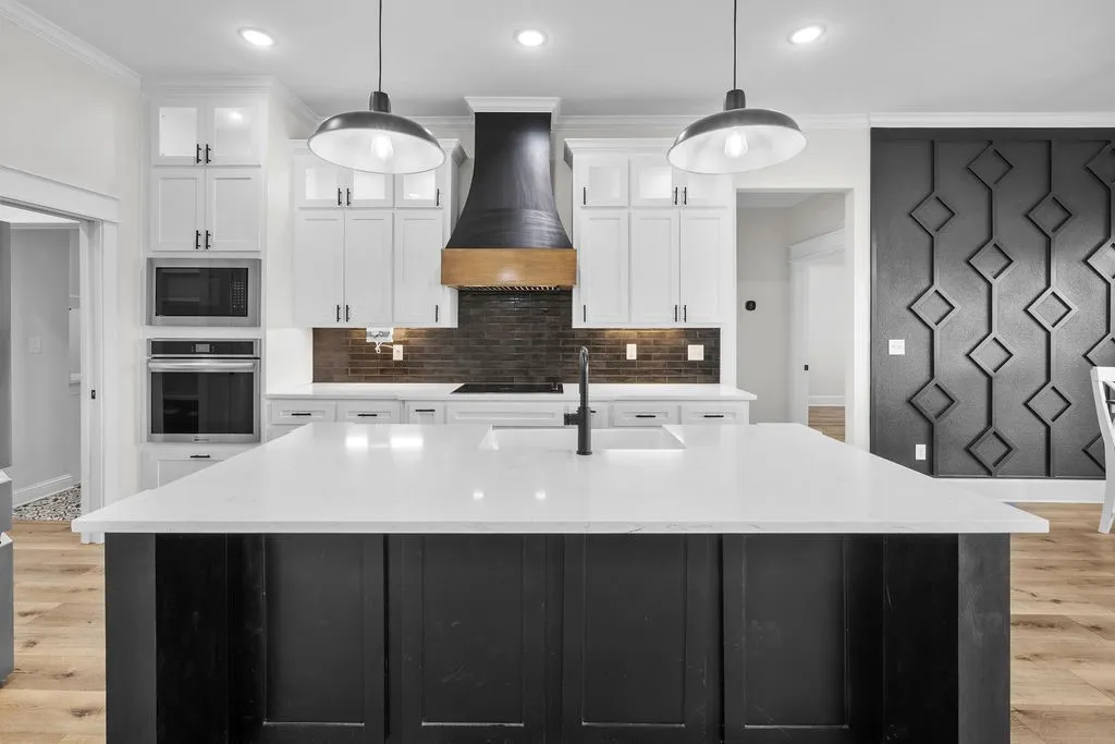 Kitchen island with a white countertop and integrated sink, pendant lighting, built-in stainless steel appliances, white cabinetry, and a dark tile backsplash