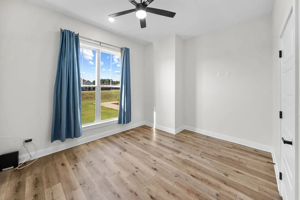 Wood-finish plank flooring, ceiling fan with integrated lighting, large window with white trim, and white paneled door with black hardware