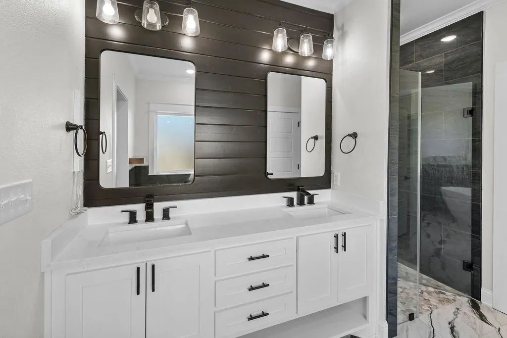 Dual vanity with white cabinetry and integrated sinks, matte black fixtures, dark wood-finish accent wall, and a separate glass-enclosed shower with tiled walls and floor