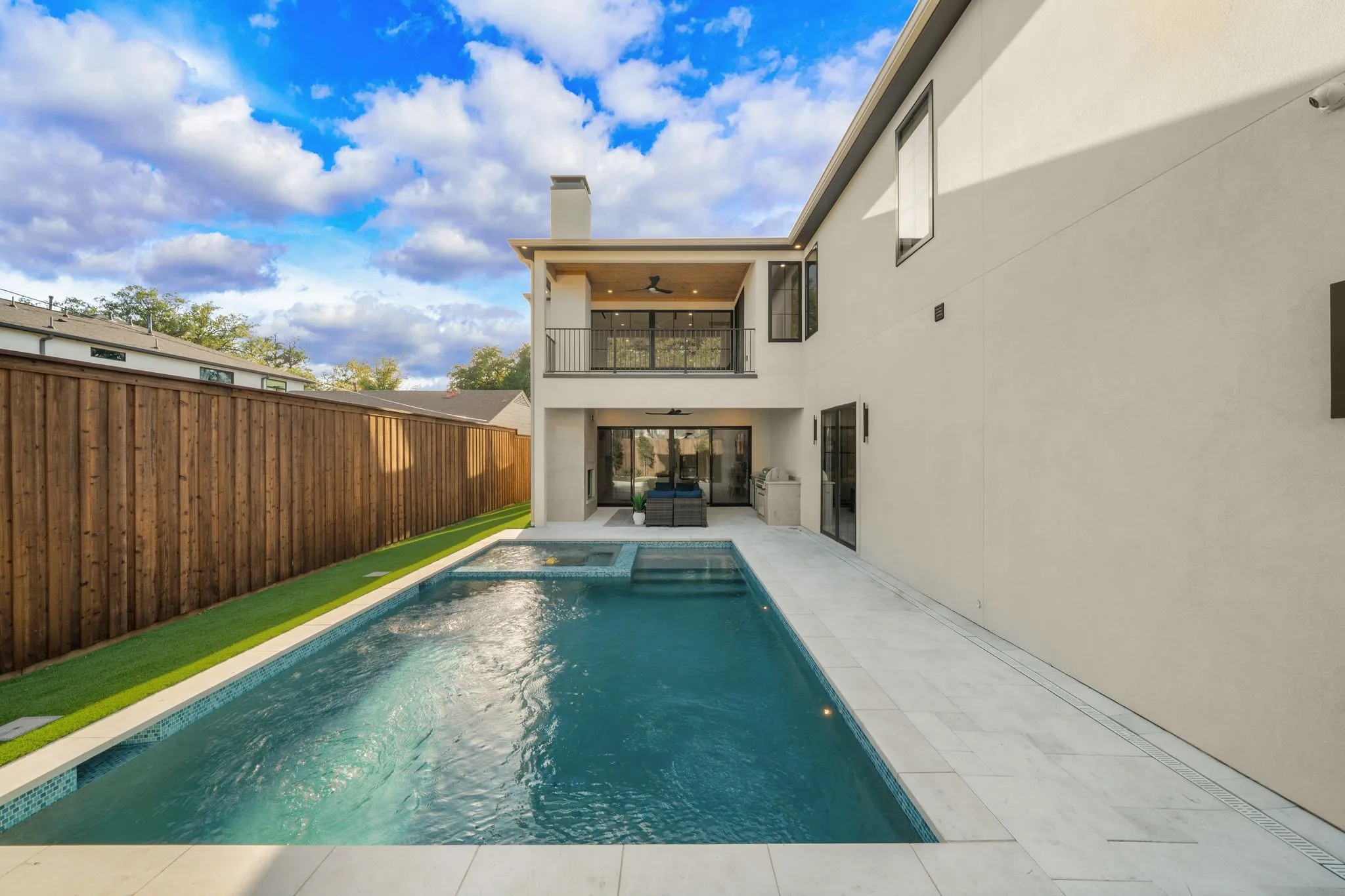 Outdoor living space featuring a rectangular pool with an integrated spa, expansive stone decking, a ground-level patio, and an upper-level covered balcony with a glass sliding door