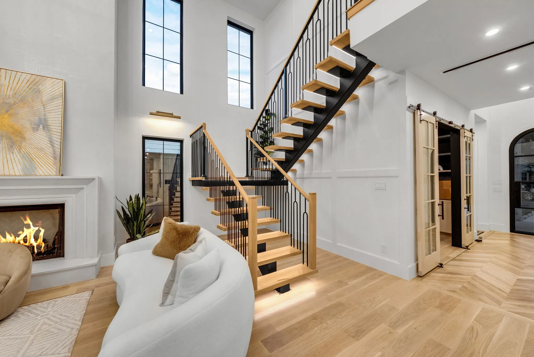 Two-story living area featuring a fireplace with a white mantel, chevron wood-finish flooring, and a floating staircase with wood treads and black metal balusters