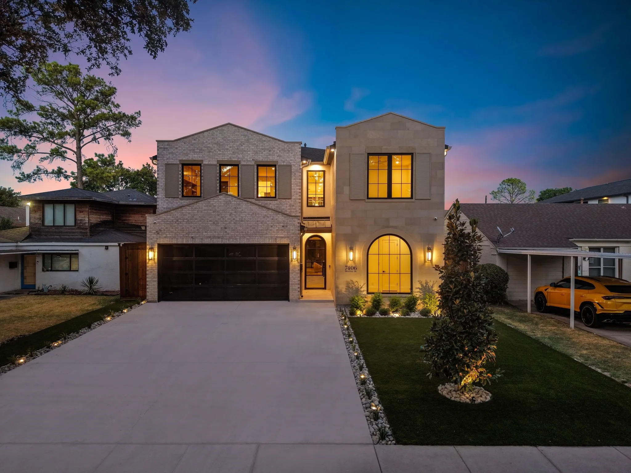 Brick and stone facade, arched entryway, manicured artificial turf, paver driveway, and exterior uplighting
