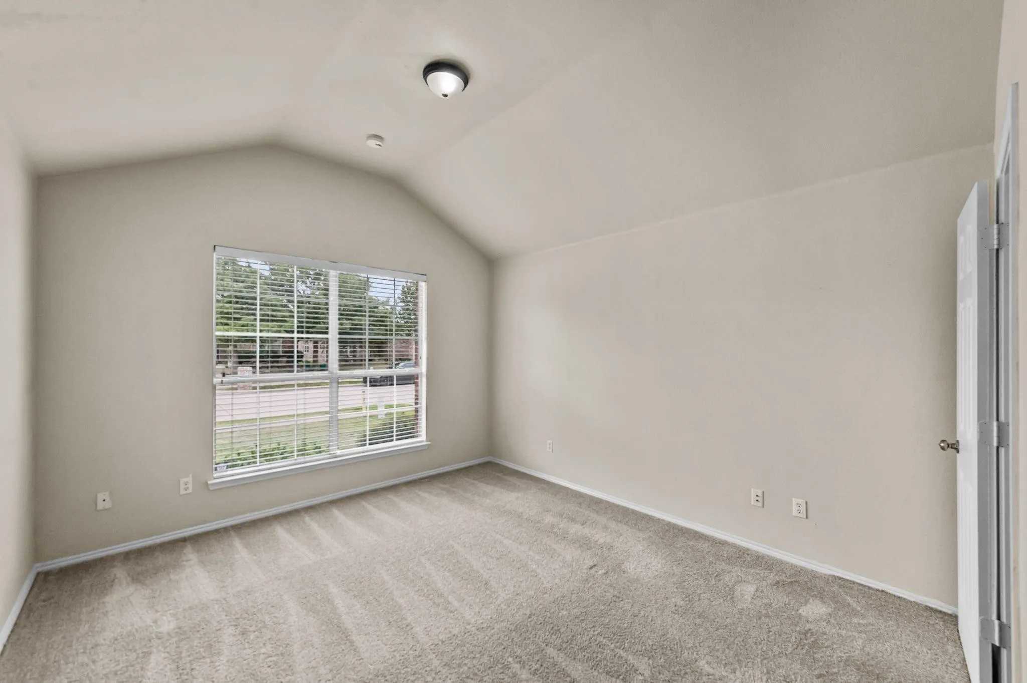 Vaulted ceiling, ceiling-mounted light fixture, neutral carpeting, window with blinds, and white trim