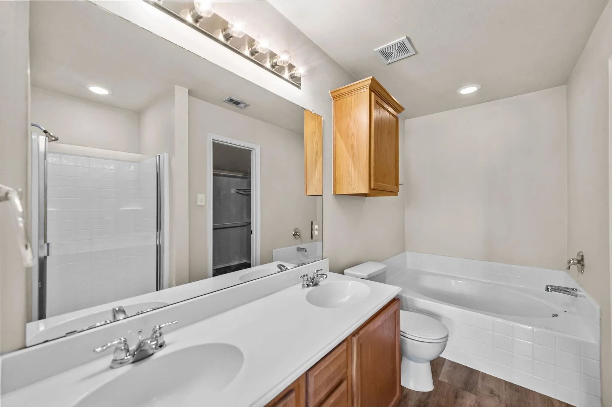 Dual vanity with integrated sinks, full-width mirror, wood-finish cabinetry, built-in soaking tub, and separate shower with glass enclosure