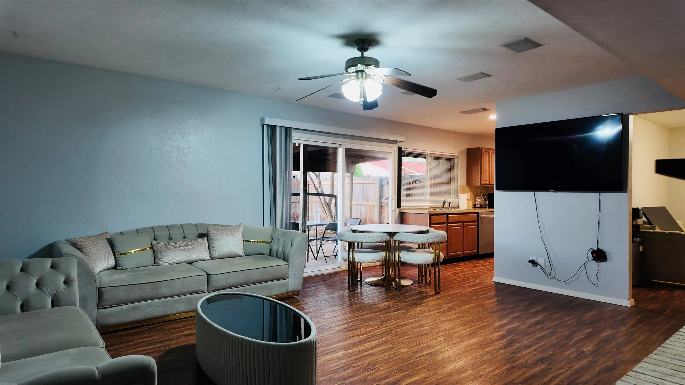 Wood-finish flooring throughout, ceiling fan with integrated lighting, sliding glass door to exterior, kitchen area with wood cabinetry and stainless steel appliances, and built-in wall partition