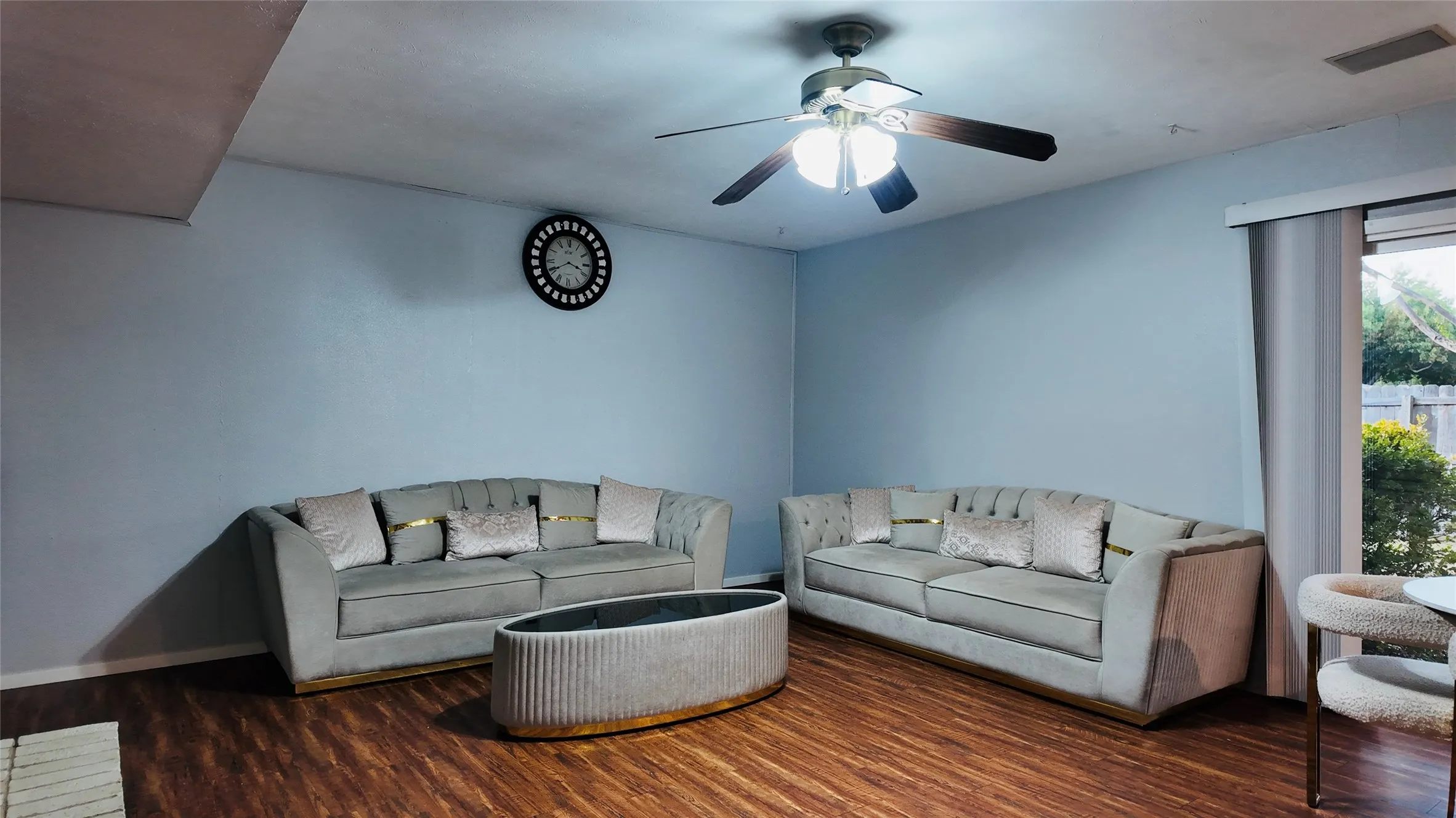 Wood-finish flooring, ceiling fan with integrated lighting, light blue wall paint, and a large window with vertical blinds