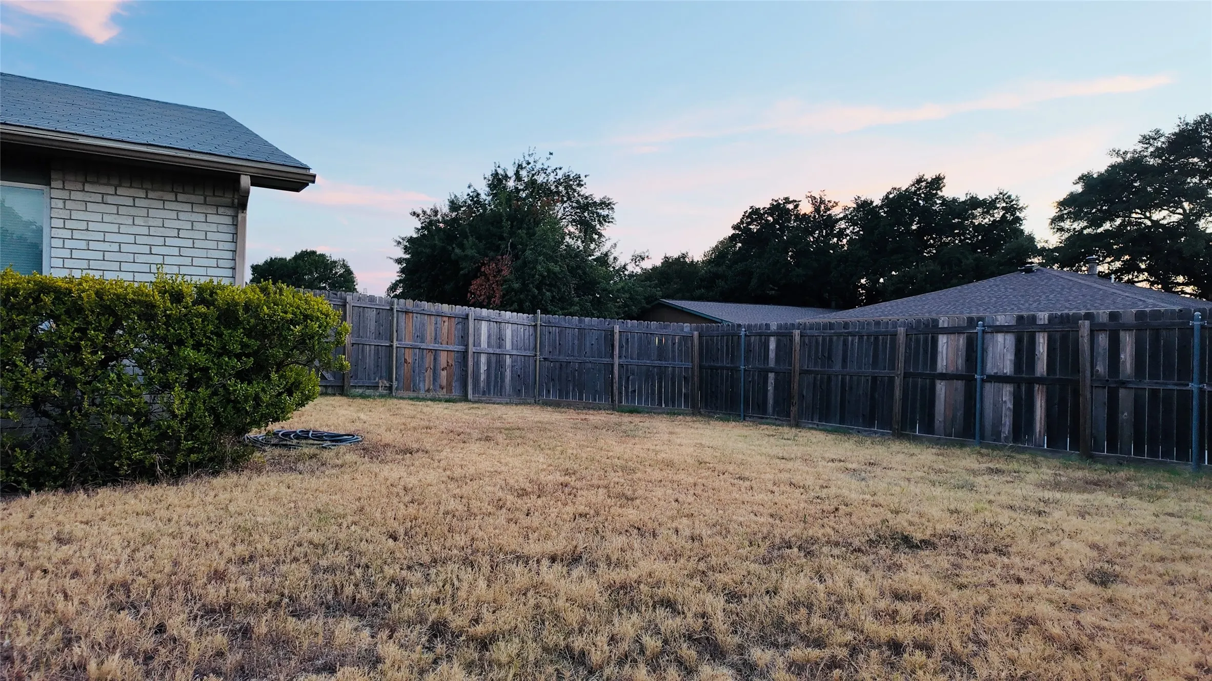 Fenced backyard with mature perimeter trees, brick exterior, and a shingled roofline