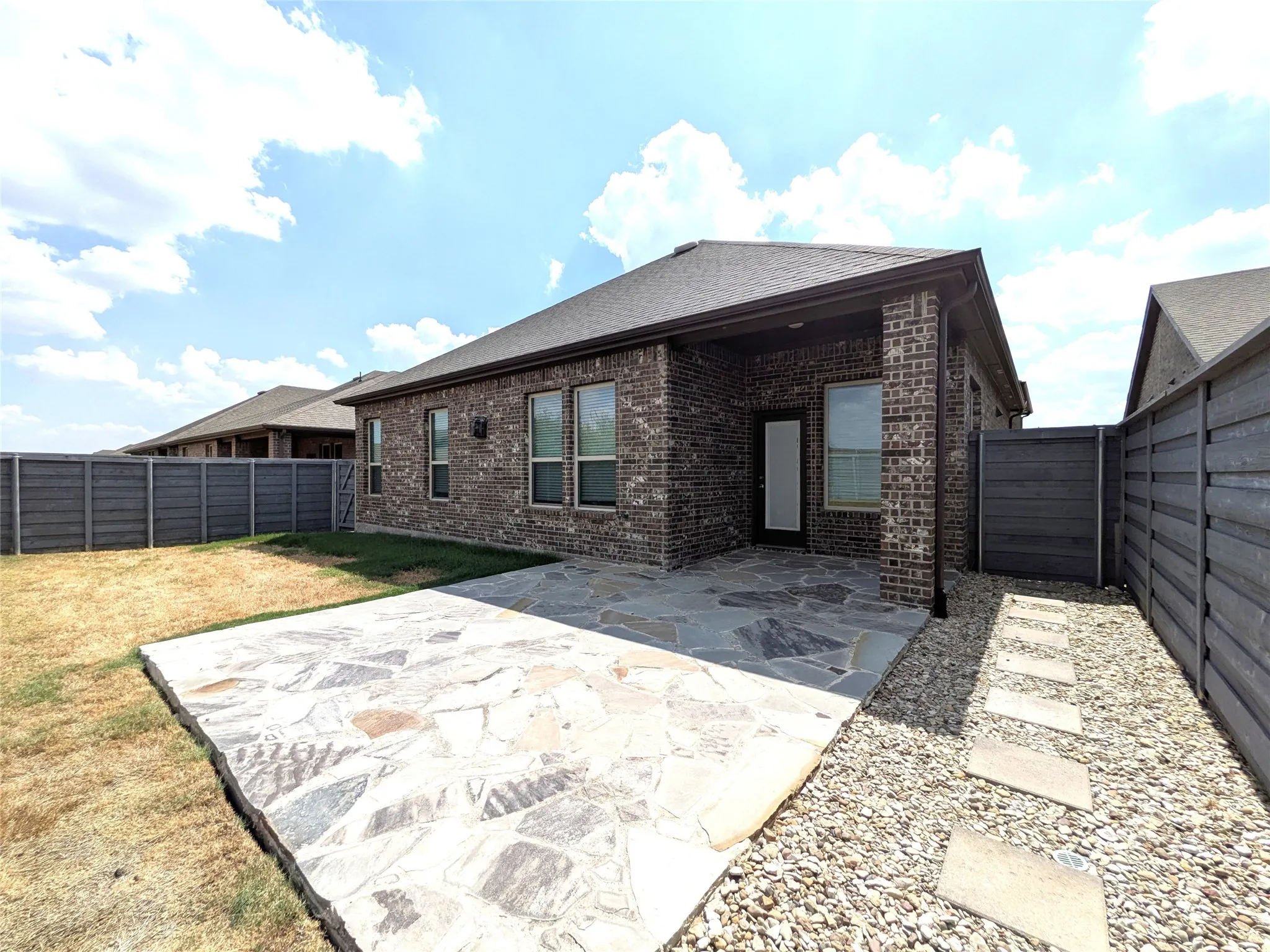 Brick exterior with dark window frames, an irregular flagstone patio, a gravel pathway with stepping stones, and a horizontal slatted privacy fence