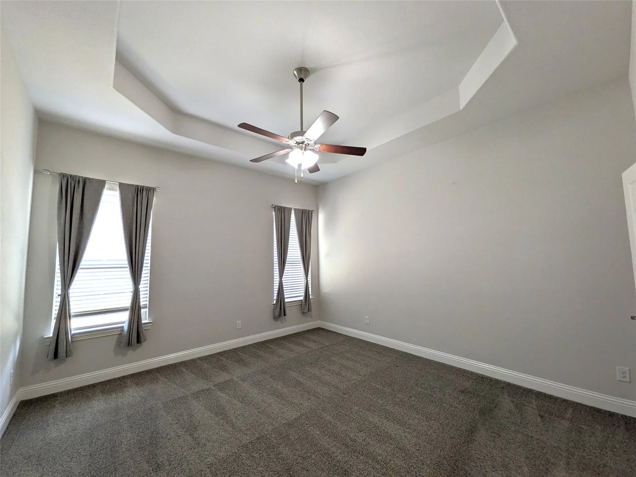 Tray ceiling, ceiling fan with light, two windows with blinds, light gray wall finish, and gray carpeted flooring