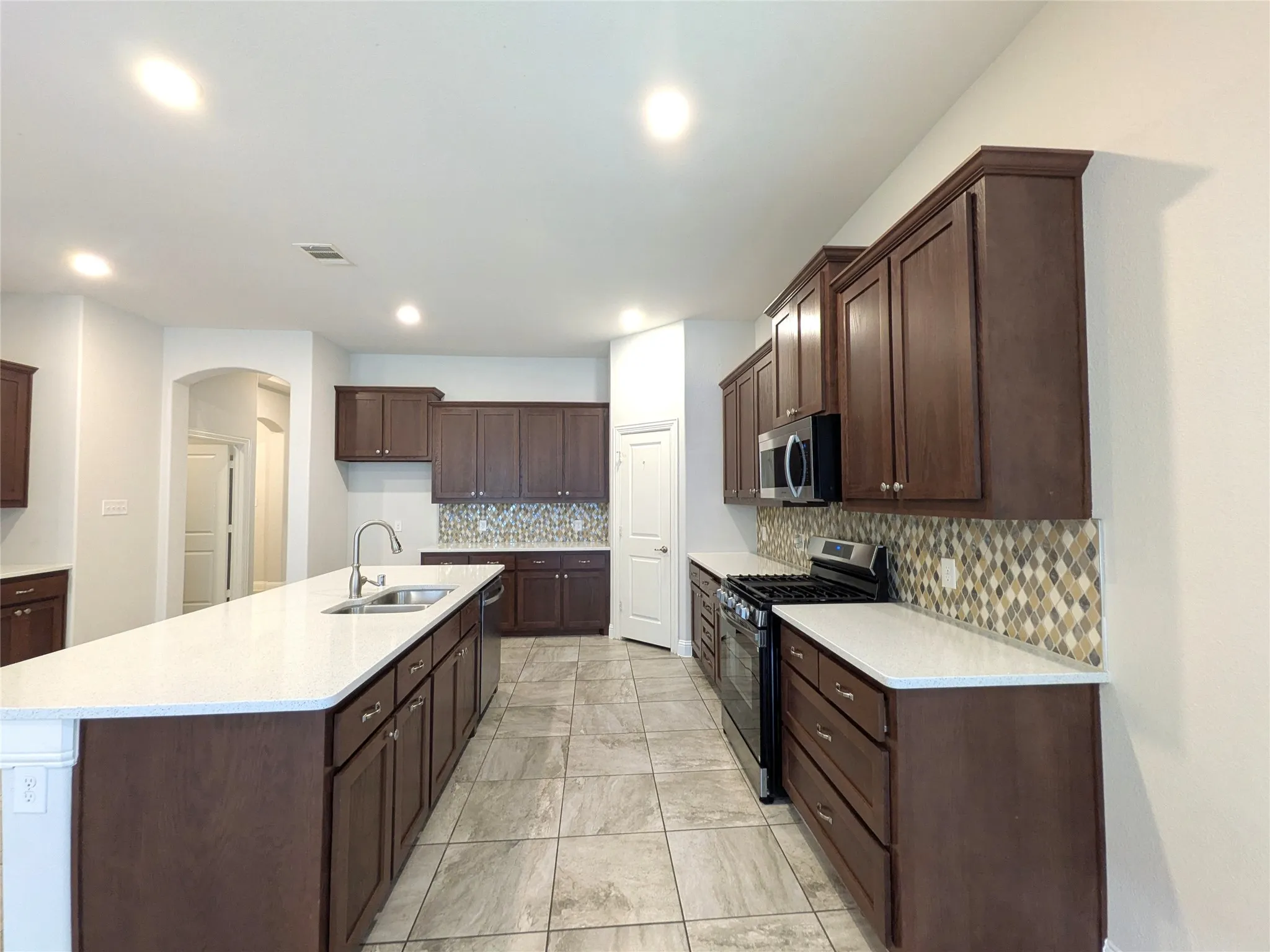 Spacious kitchen featuring dark wood-finish cabinetry, light-toned countertops, full-height decorative tile backsplash, stainless steel appliances, and large format tile flooring