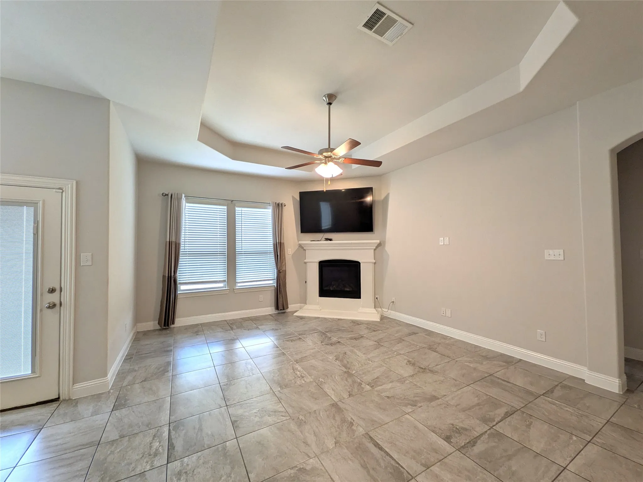 Tray ceiling with a ceiling fan, tiled flooring, corner fireplace, and multiple windows with blinds