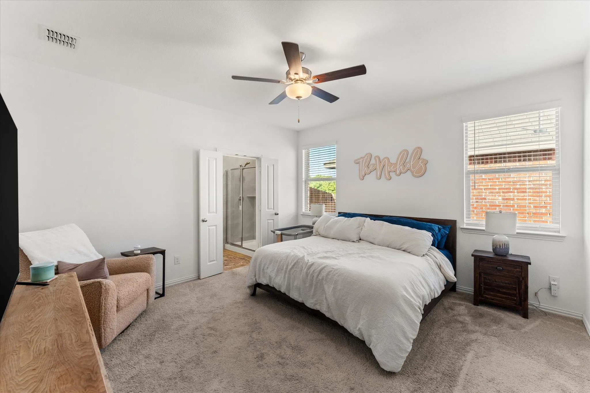 Carpeted bedroom with a ceiling fan, two windows with blinds, and dual doors opening to a bathroom featuring a glass-enclosed shower