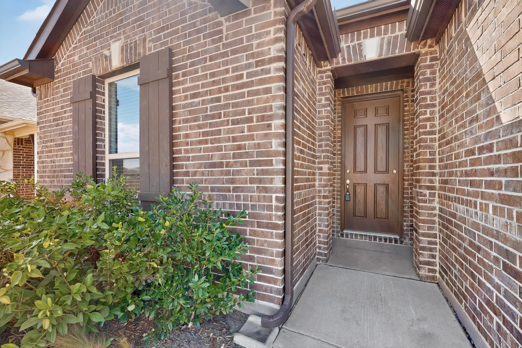 Brick exterior with a paneled entry door, window with dark shutters, and established landscaping