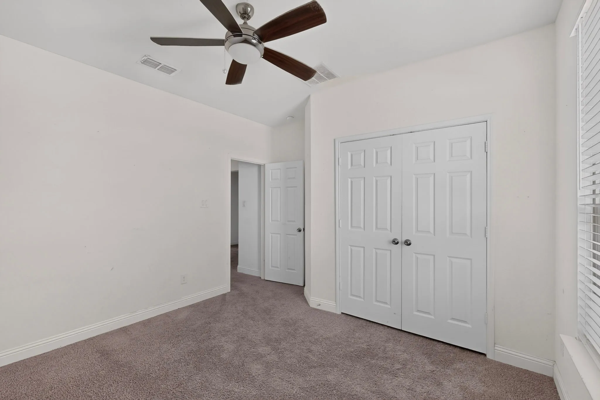 Neutral tone wall paint, textured carpet flooring, ceiling fan with wood-finish blades, and double bifold closet doors