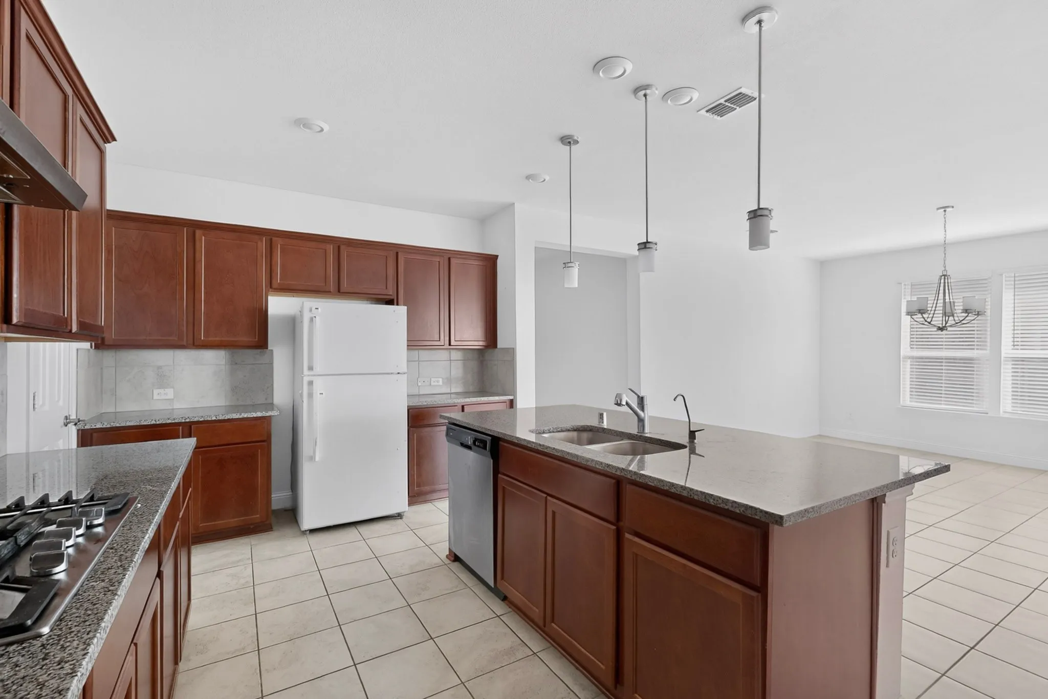 Kitchen featuring wood-finish cabinetry, granite countertops, a central island with sink, tile flooring, and pendant lighting
