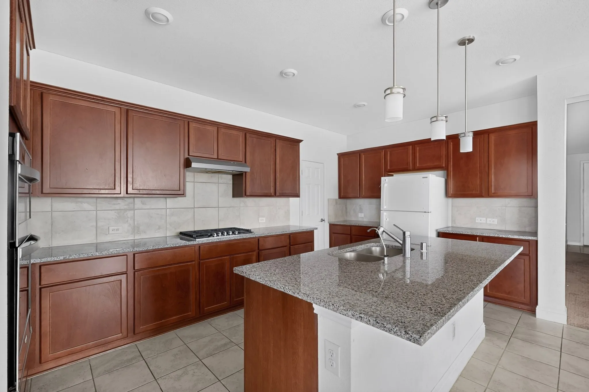 Kitchen featuring wood-finish cabinetry, granite countertops, tile backsplash, a gas range with a stainless steel hood, and a kitchen island with an integrated sink and pendant lighting