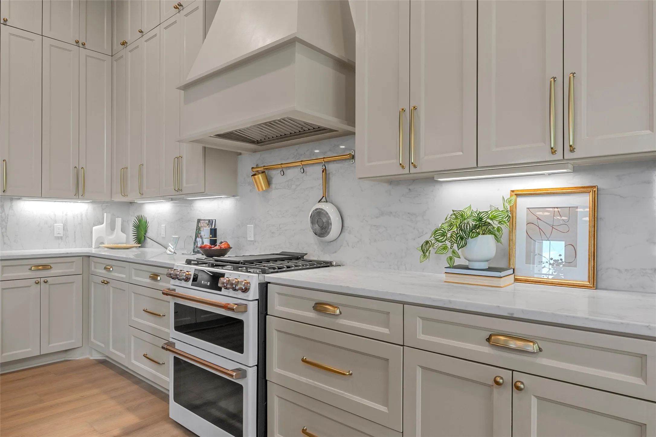 Light-toned cabinetry with gold-toned hardware, stone countertops, stone backsplash, under-cabinet lighting, and a double oven range with a coordinating vent hood
