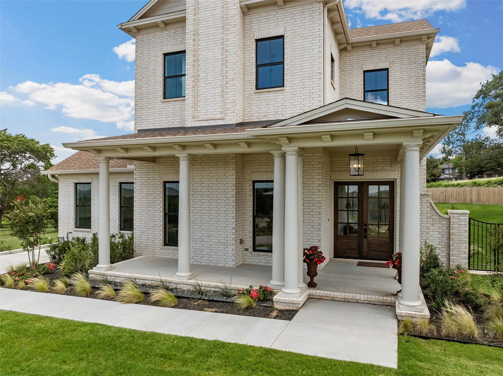 White brick exterior with black-framed windows, a covered entry porch with columns, dark wood-finish double entry doors, and mature landscaping