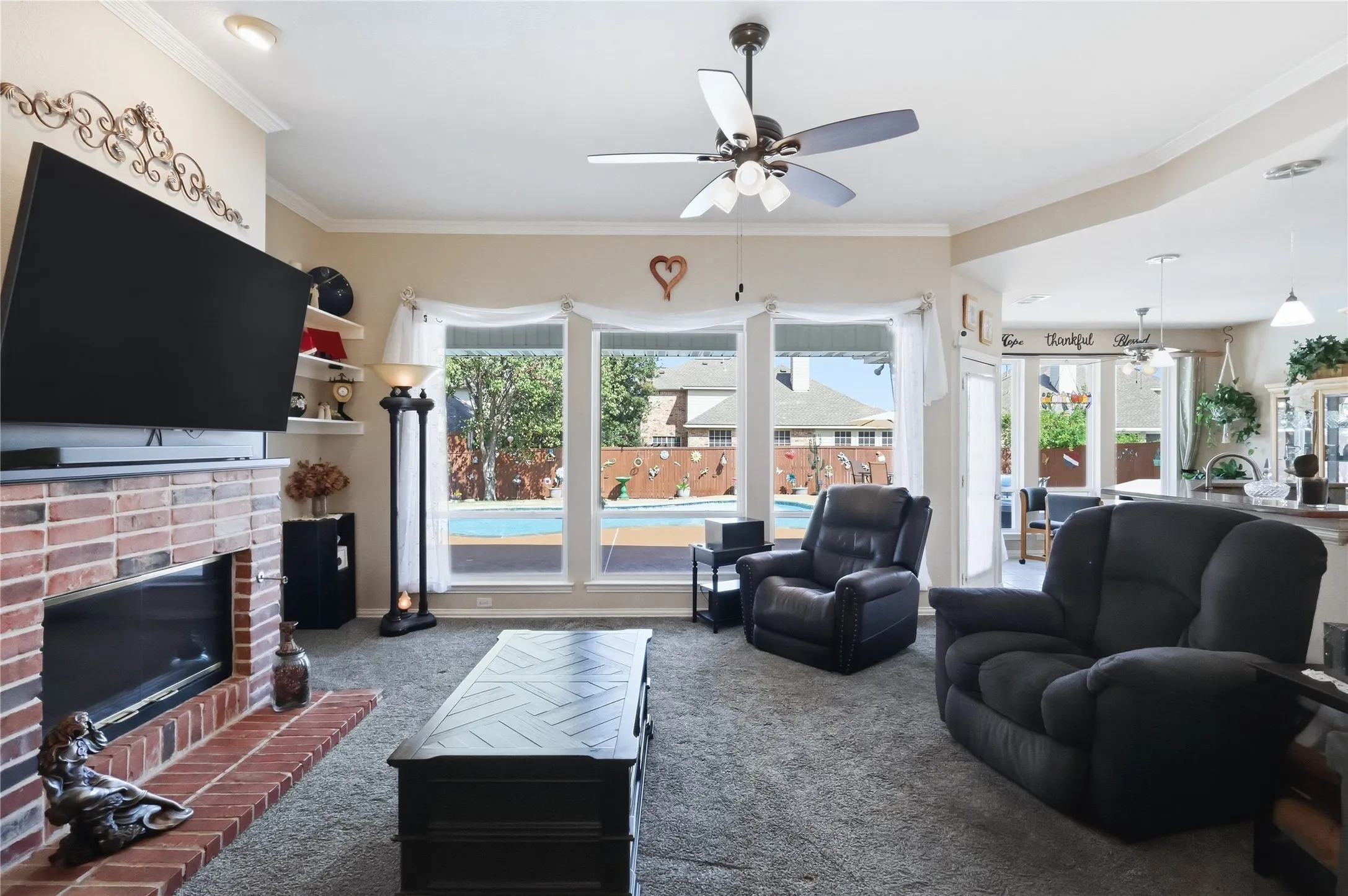 Living area with a brick fireplace, built-in shelving, ceiling fan, and large windows overlooking a backyard pool